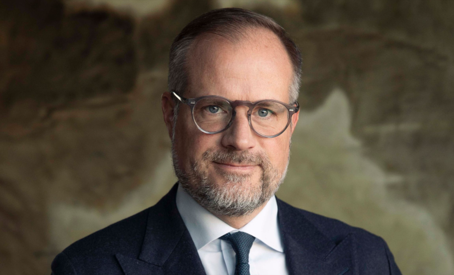 Porträttfoto av en man med glasögon och skägg, klädd i mörkblå kostym och mörkblå slips mot en suddig bakgrund.