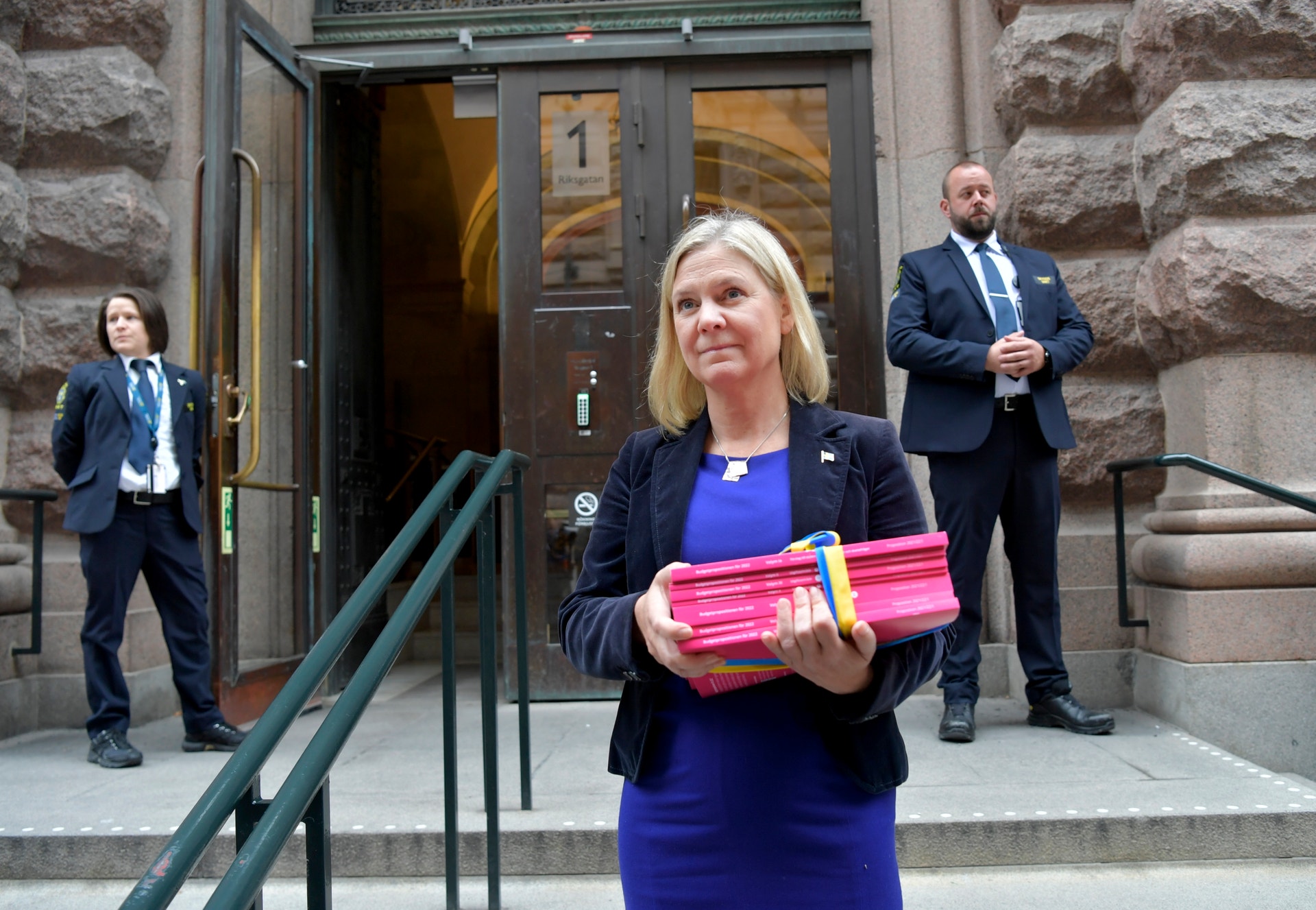 En kvinna i blå kostym står framför en stenbygnad och håller ett paket inslaget i rosa papper. I bakgrunden syns två män i kostym vid byggnaden.