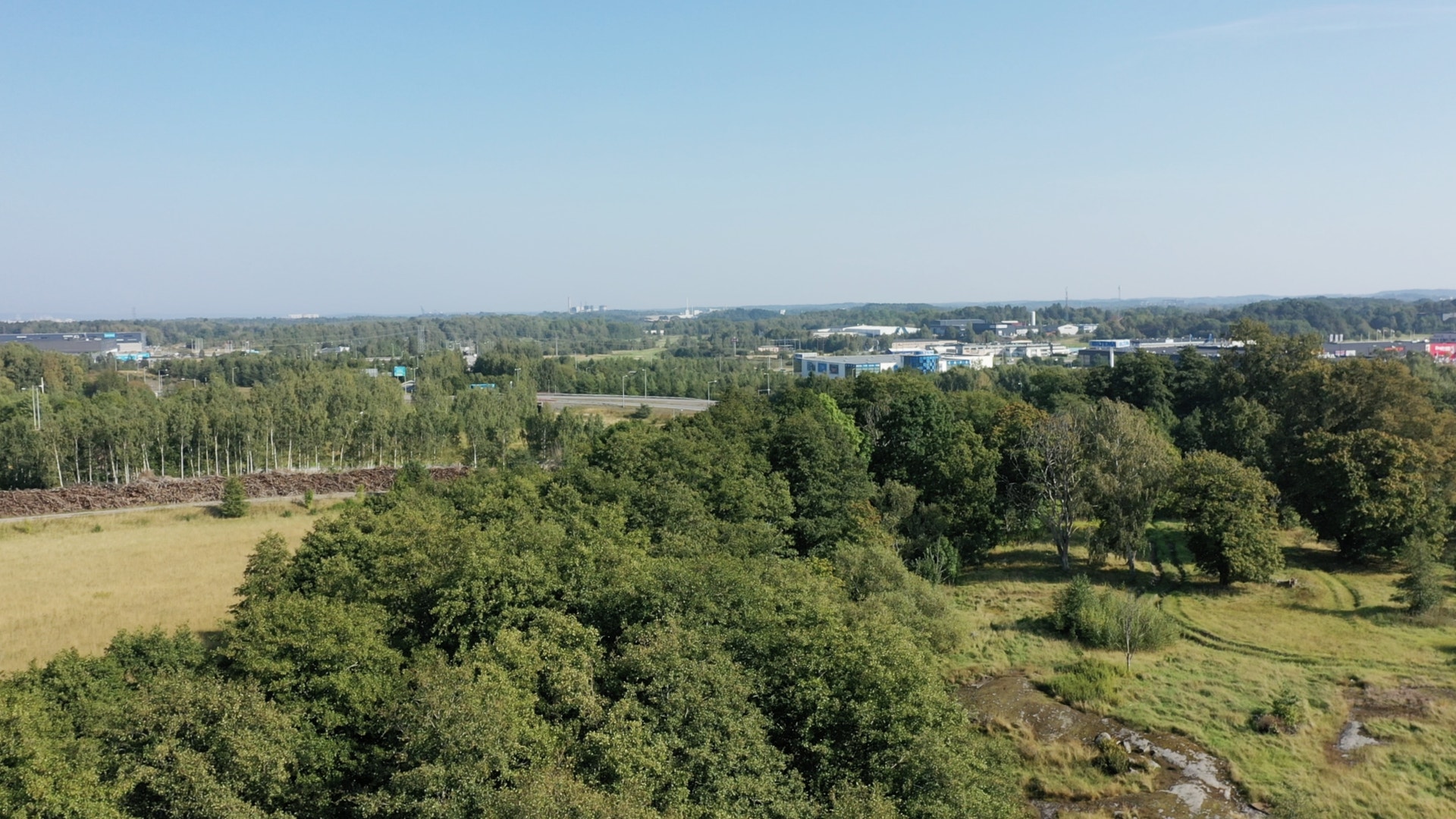 Flygbild över ett grönt landskap med skog och öppna gräsytor. I fjärran syns låga industribyggnader under en klarblå himmel.