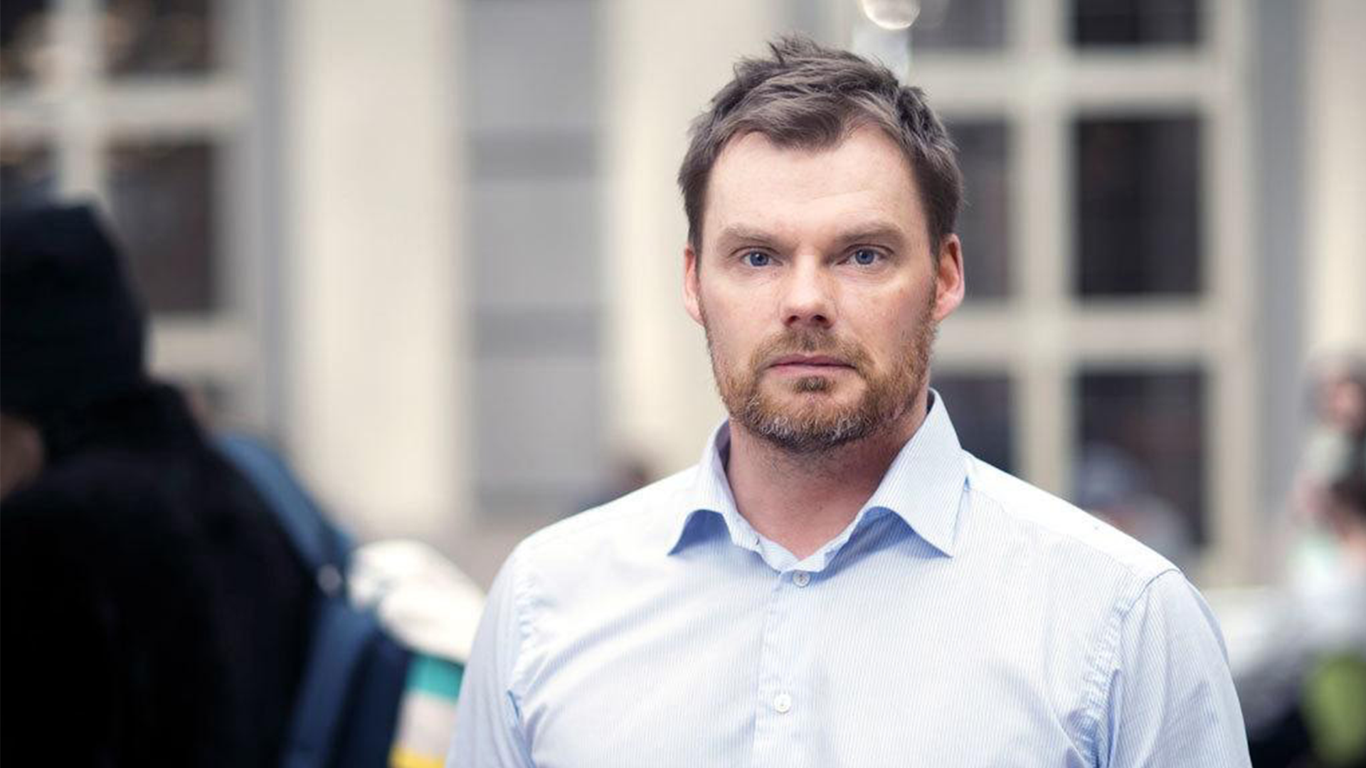 Rickard Sandberg poserar vid Handelshögskolan. Han bär blå skjorta och tittar seriöst in i kameran.