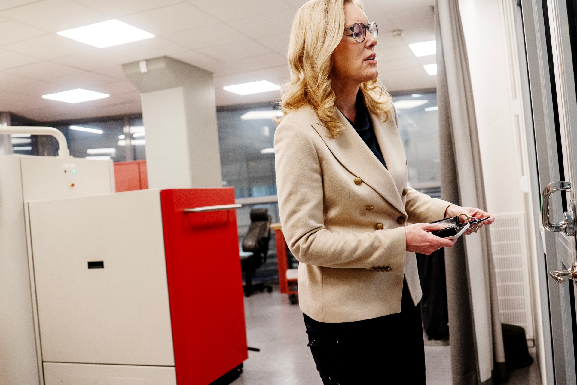 Kvinna i beige kavaj står i ett modernt kontor och håller en smartphone