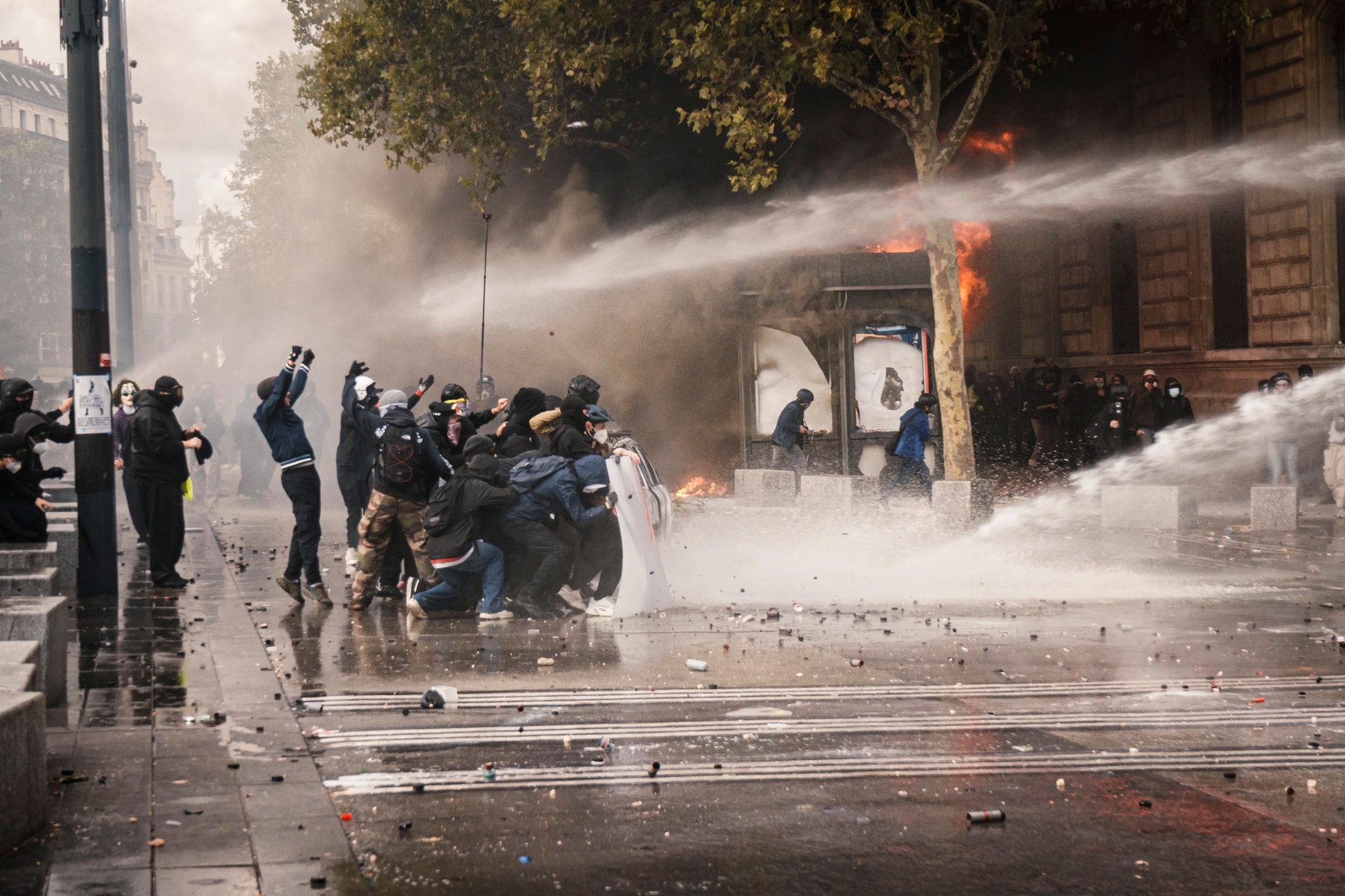 Maskerade demonstranter tar skydd tillsammans på en gata medan kraftiga vattenstrålar sprutar över dem.