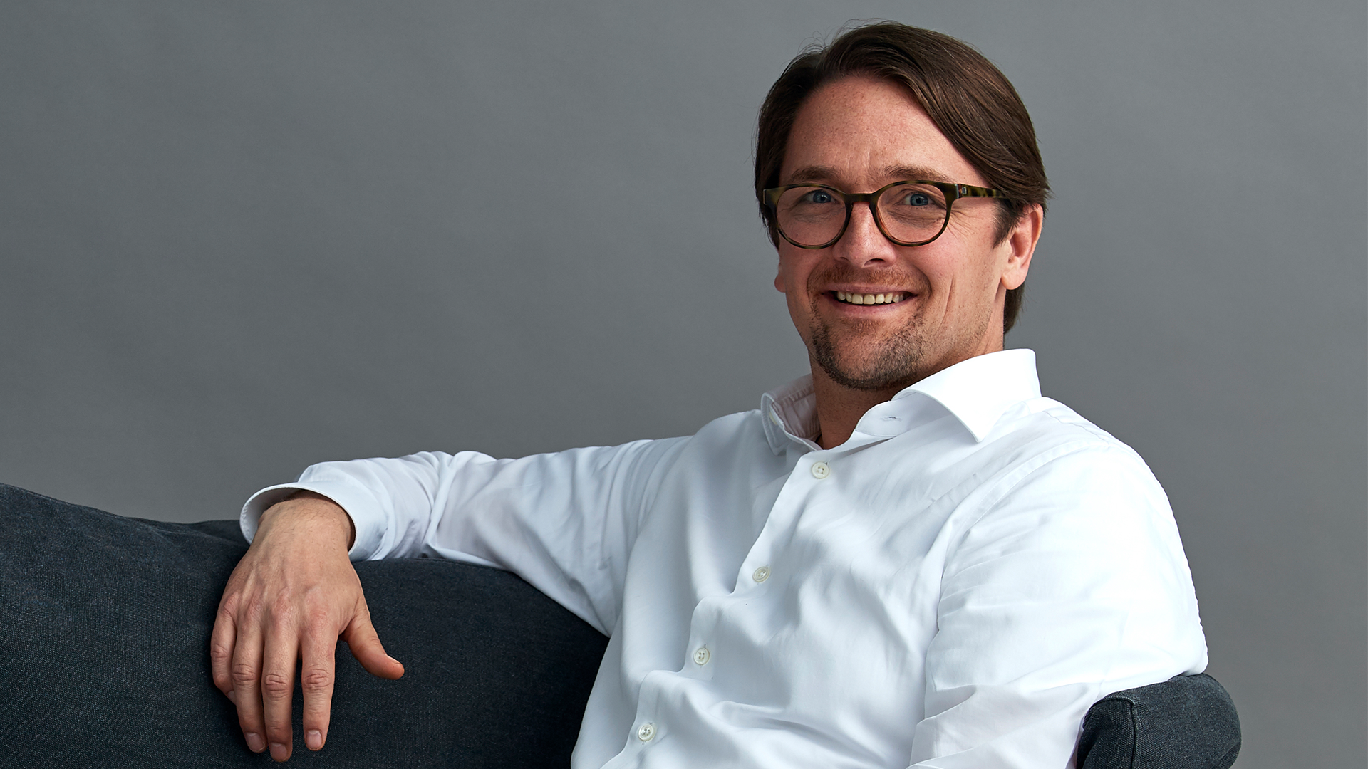 Porträtt av en man med glasögon och vit skjorta som sitter på en mörkgrå soffa och ler mot kameran.