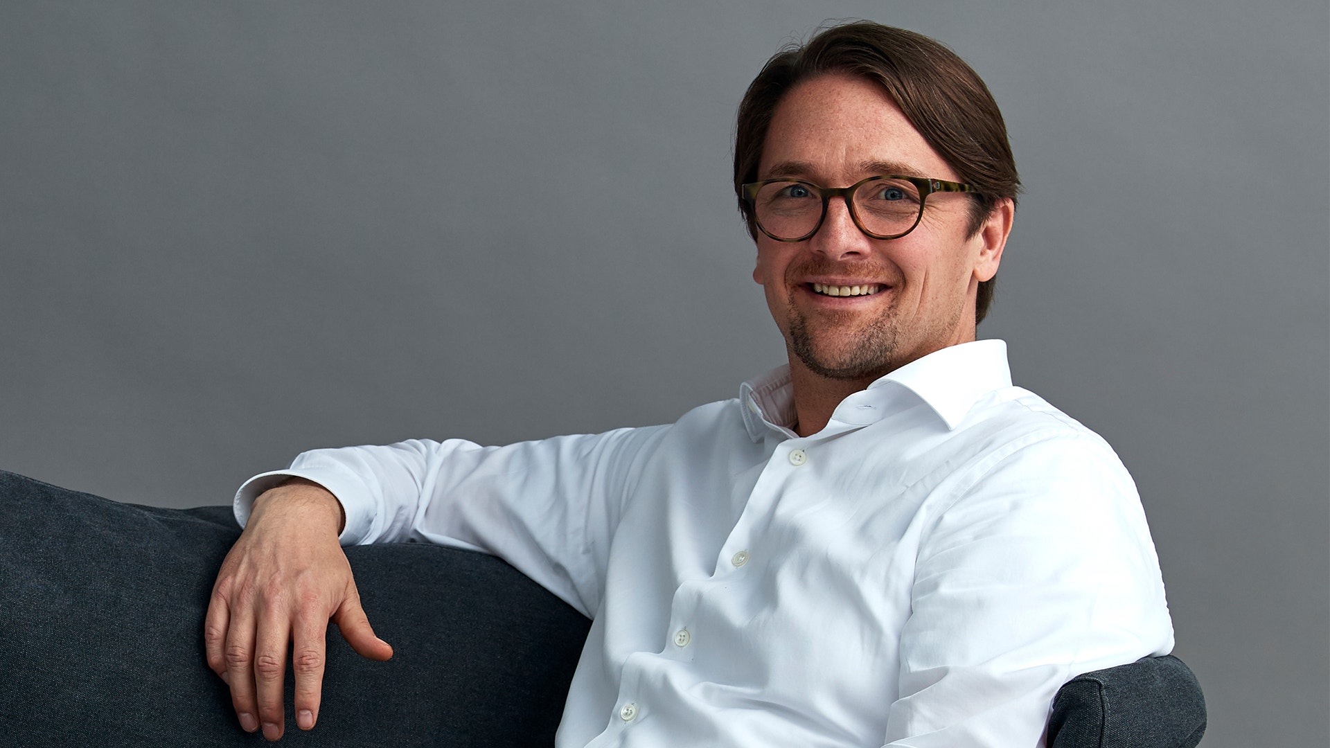 Porträtt av en man med glasögon och vit skjorta som sitter på en mörkgrå soffa och ler mot kameran.
