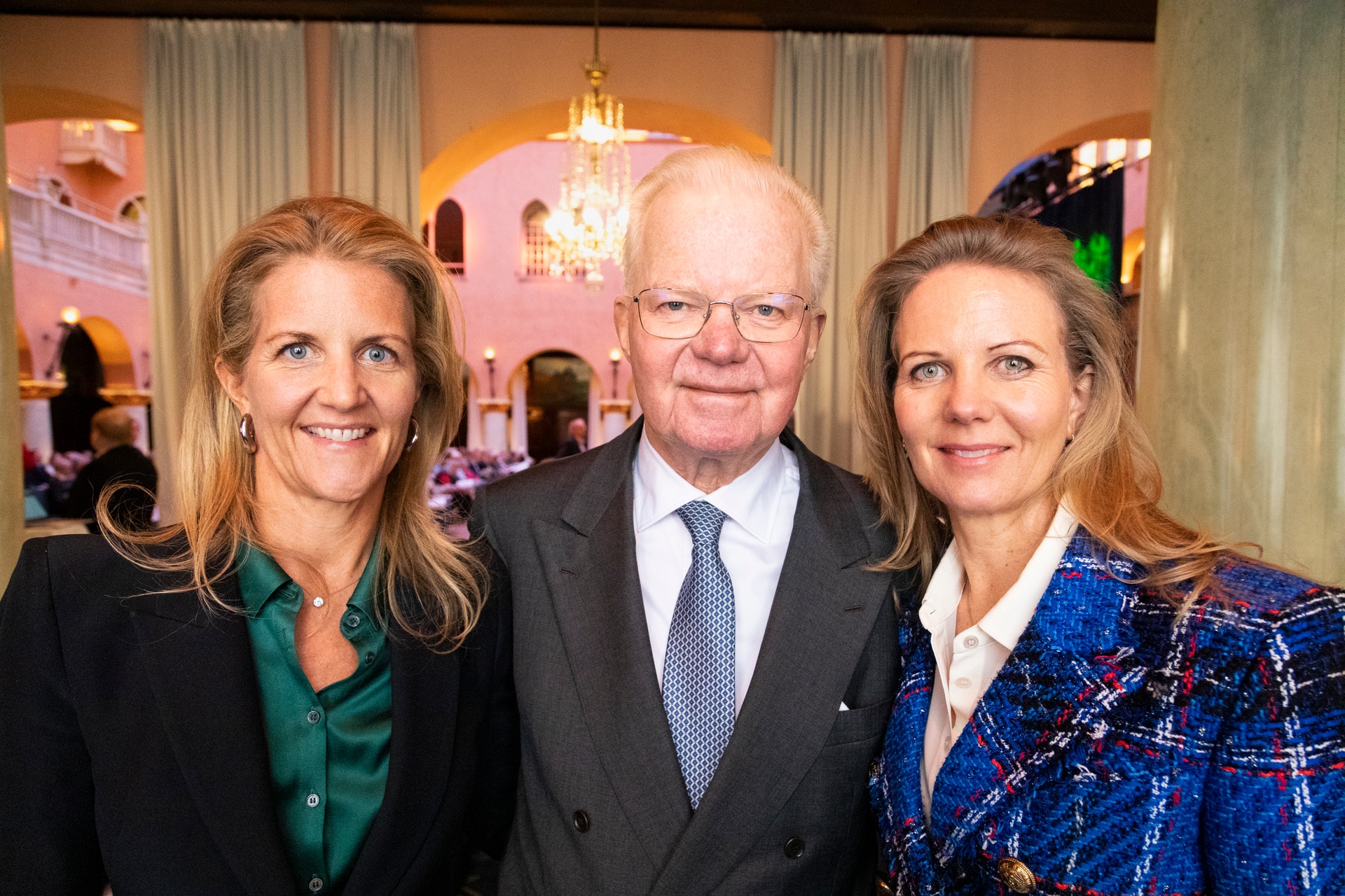 Tre personer poserar tillsammans vid ett formellt evenemang. En äldre man i kostym står i mitten mellan två kvinnor i elegant kläder. I bakgrunden syns en elegant sal med kristallkrona.
