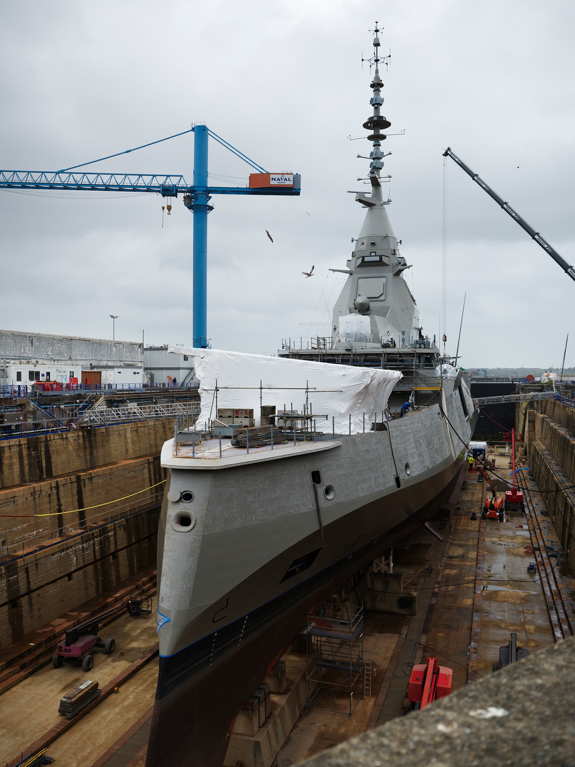 Gråmålat marinfartyg i skeppsdocka omgivet av byggnadsställningar och en stor blå kran.