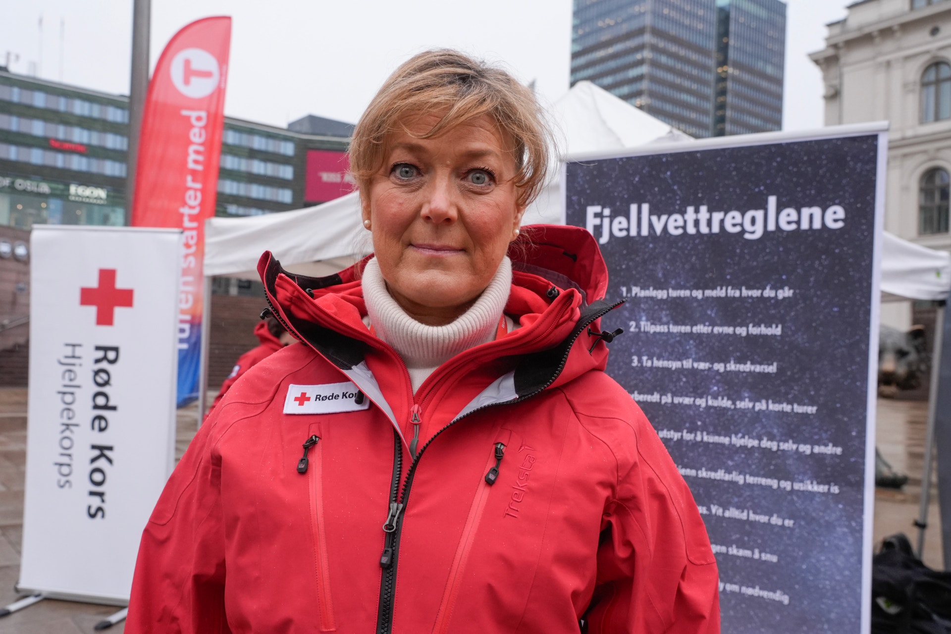 Kvinna i röd jacka från Röda Korset står utomhus framför en informationsstand.