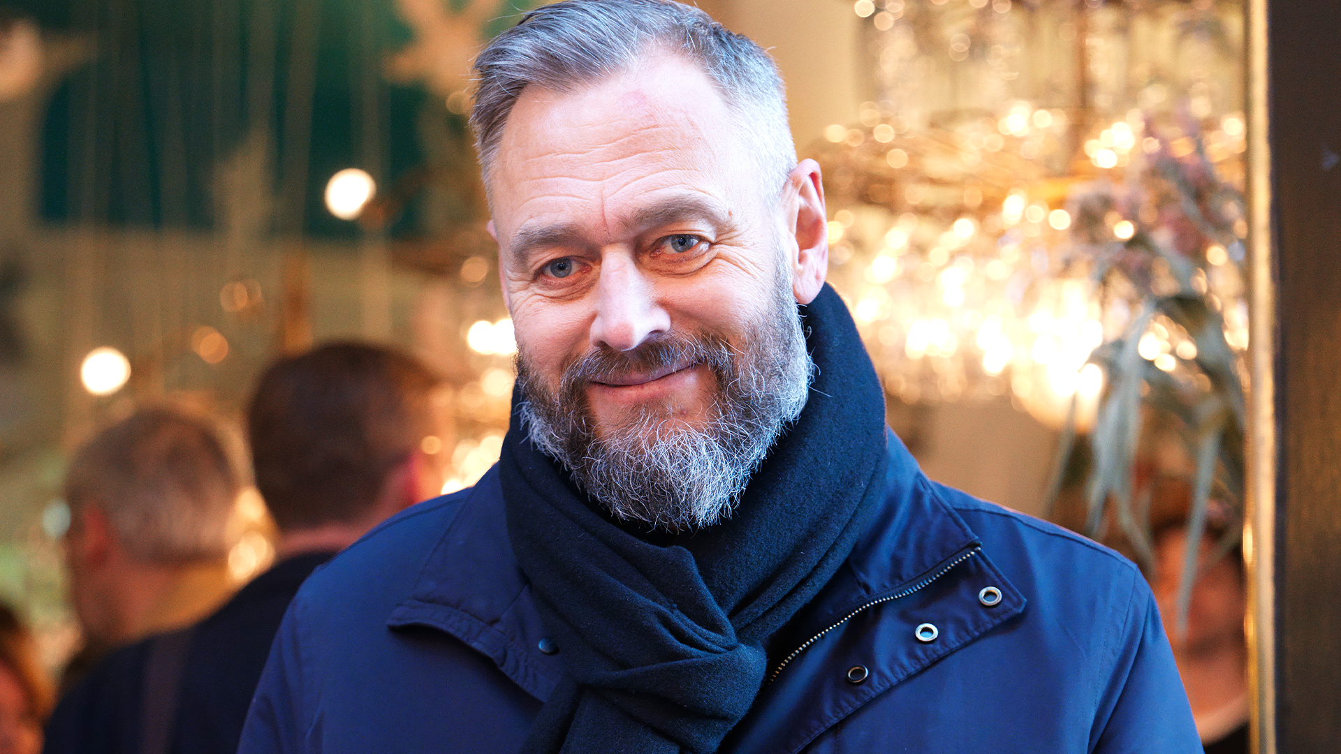 Porträtt av en man med skägg och glasögon i mörkblå jacka framför en suddig festlig bakgrund med varmt ljus.