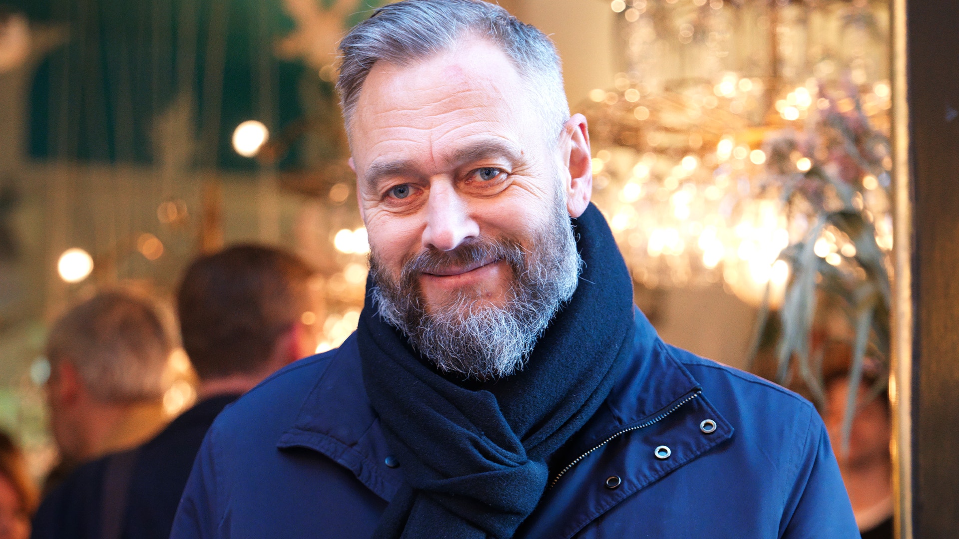 Porträtt av en man med skägg och glasögon i mörkblå jacka framför en suddig festlig bakgrund med varmt ljus.