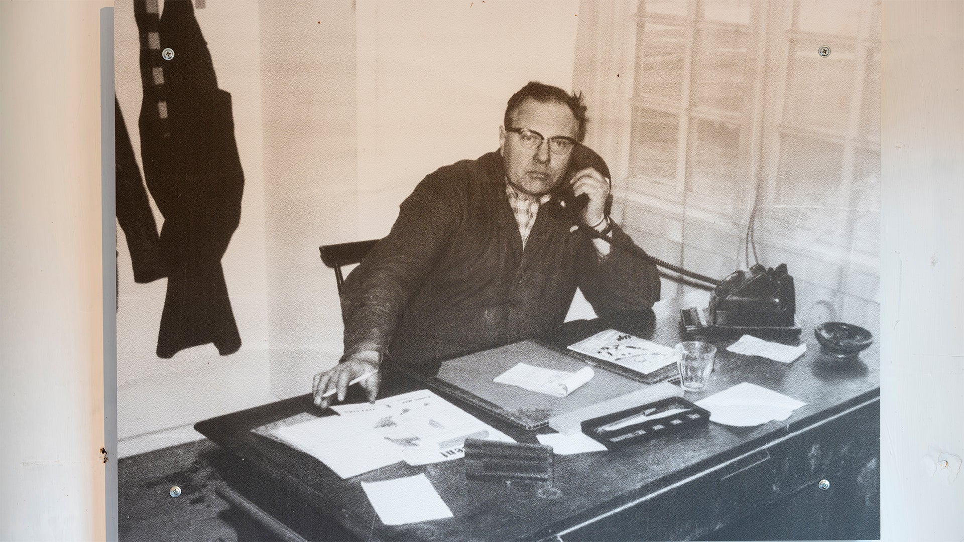 Svartvit fotografi av en man med glasögon som sitter vid ett skrivbord på ett kontor. Han är klädd i mörk kostym och arbetar omgiven av papper, skrivmaskin och telefon.
