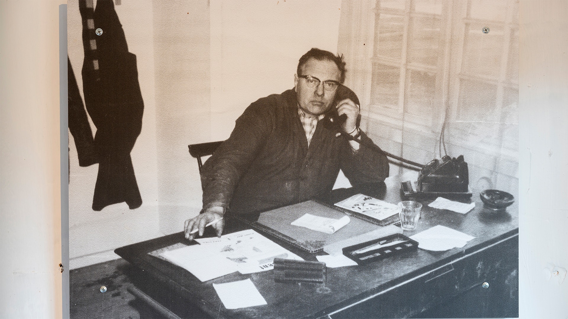 Svartvit fotografi av en man med glasögon som sitter vid ett skrivbord på ett kontor. Han är klädd i mörk kostym och arbetar omgiven av papper, skrivmaskin och telefon.