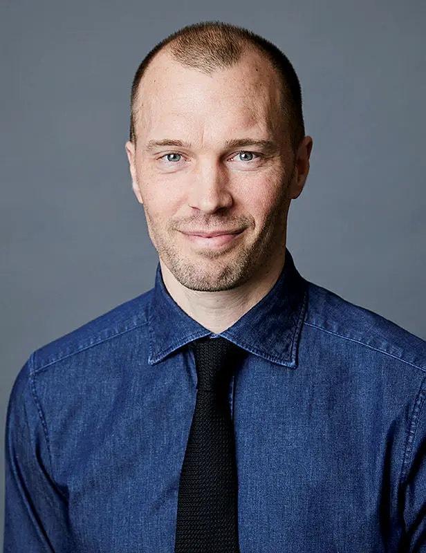 Porträtt av en man i blå jeansöverrock och svart slips mot grå bakgrund.