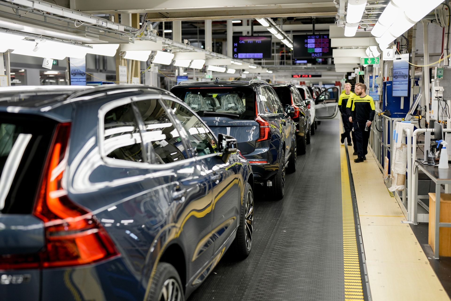 Nytillverkade bilar står på rad på en monteringslinje i en bilfabrik, medan två arbetare i gula och svarta arbetskläder går bredvid.