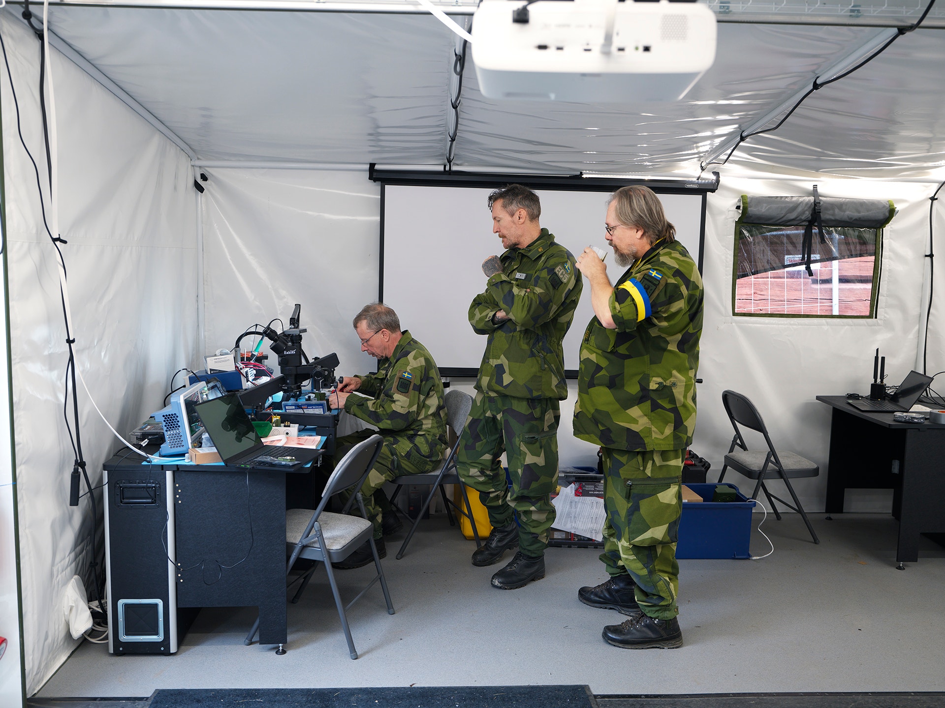 Tre personer i militäruniform arbetar med teknisk utrustning inne i ett tält.