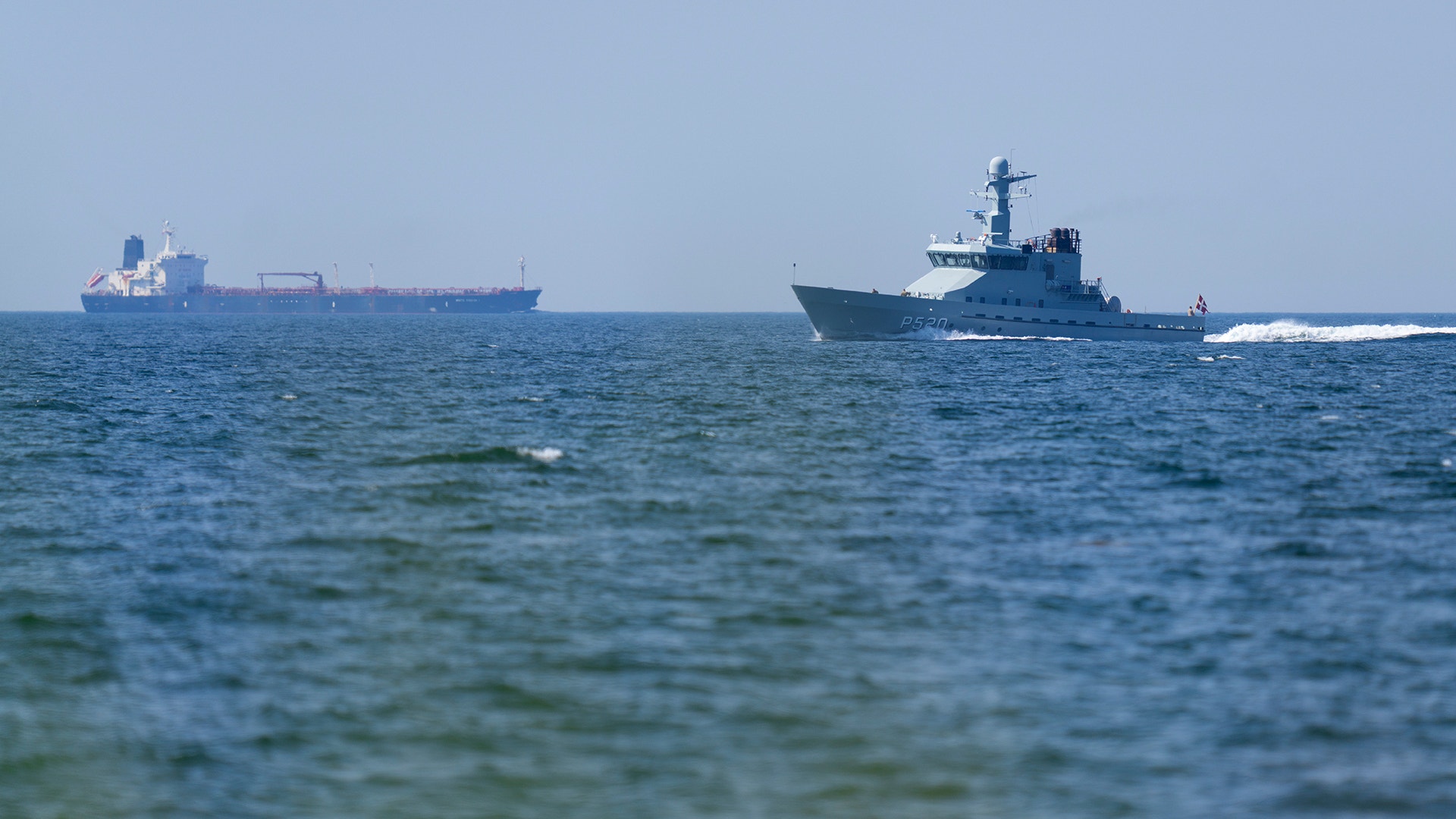 Två fartyg på havet. Ett större tankfartyg på vänster sida och ett militärt patrullfartyg på höger sida. Blått hav och klar himmel.