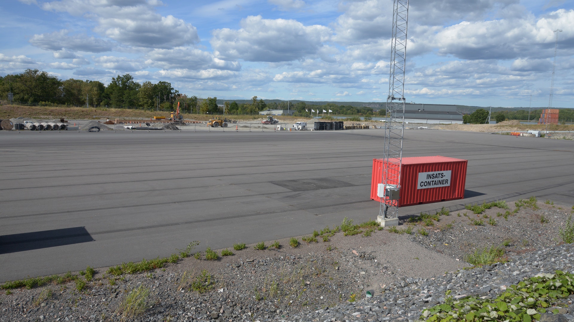 Industriområde med stor asfaltsyta, röd container och telekommunikationstorn. Träd och himmel med moln i bakgrunden.