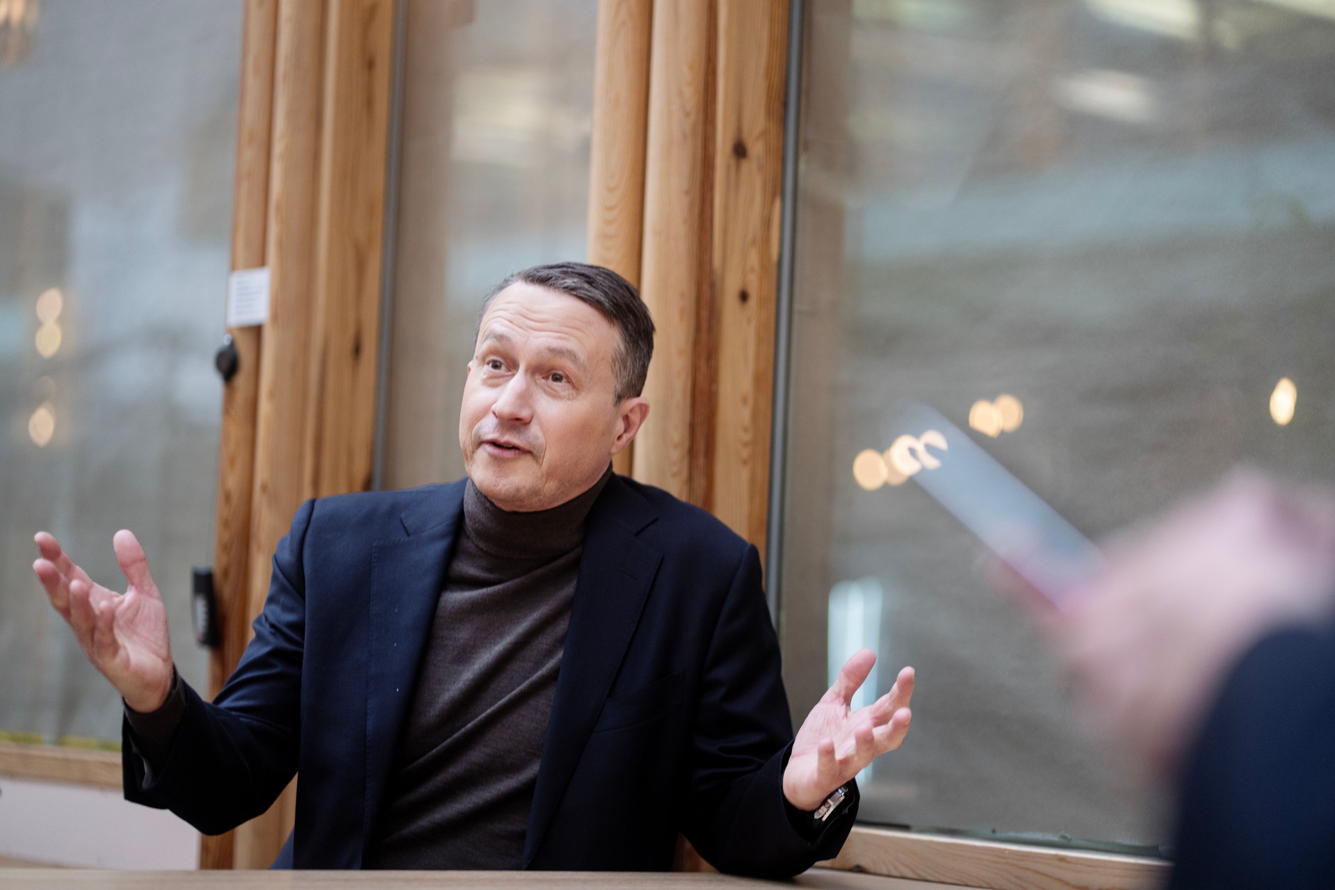 En man i mörkblå kavaj och svart tröja sitter vid ett bord och gestikulerar medan han talar.