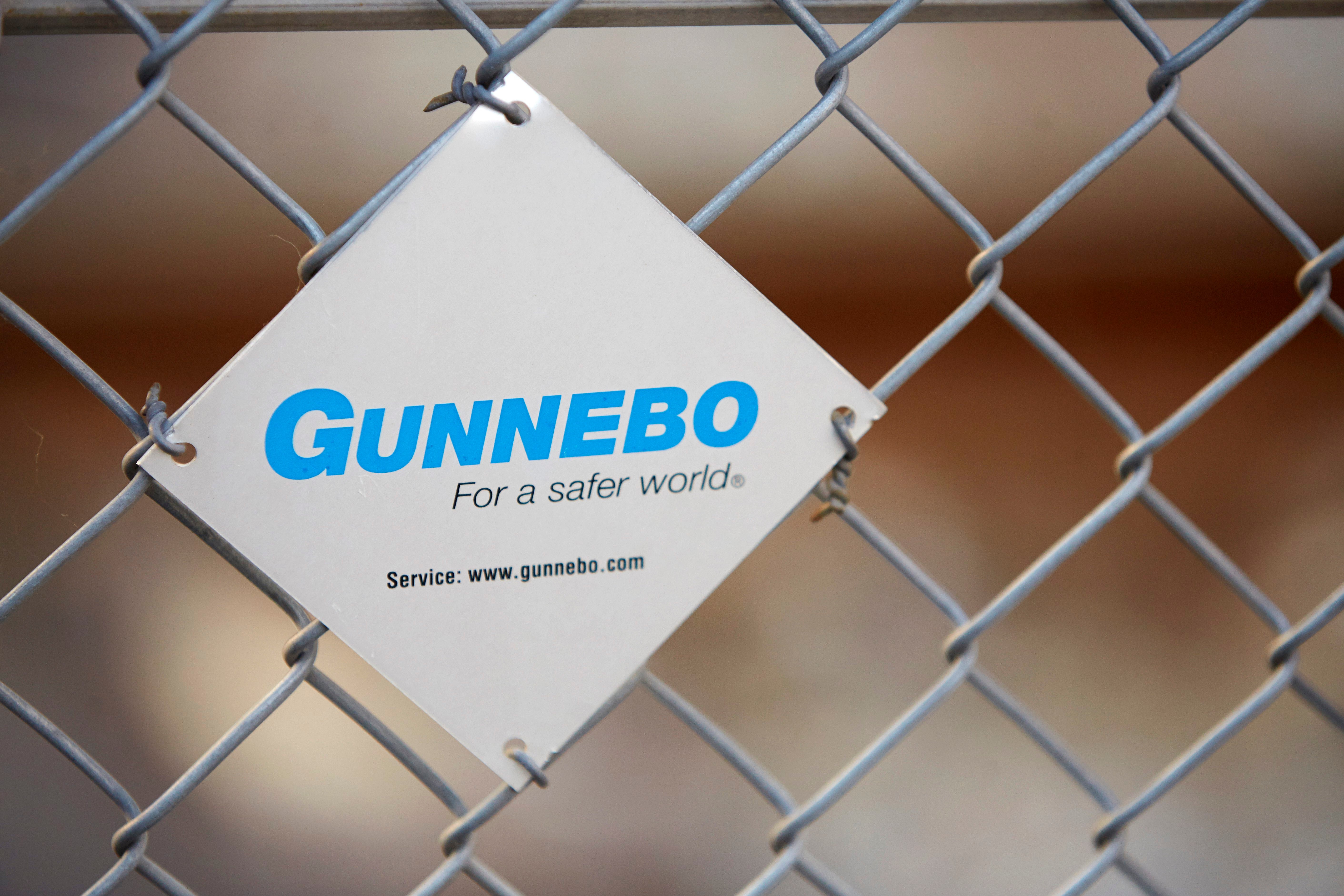 Vit skylt med blå Gunnebo-logotyp och texten 'För en säkrare värld' fäst på ett metallstängsel