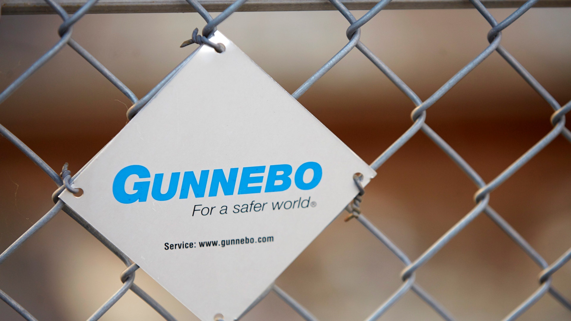 Vit skylt med blå Gunnebo-logotyp och texten 'För en säkrare värld' fäst på ett metallstängsel
