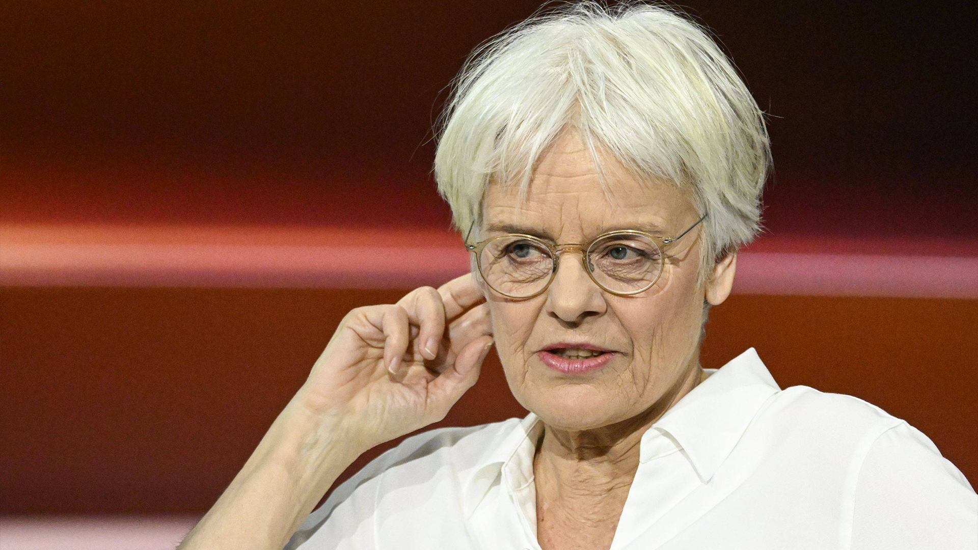 Porträtt av Ulrike Herrmann med vitt hår och glasögon, klädd i en vit skjorta. Hon lutar sitt huvud mot sin hand i en eftertänksam pose mot en rödbrun bakgrund.