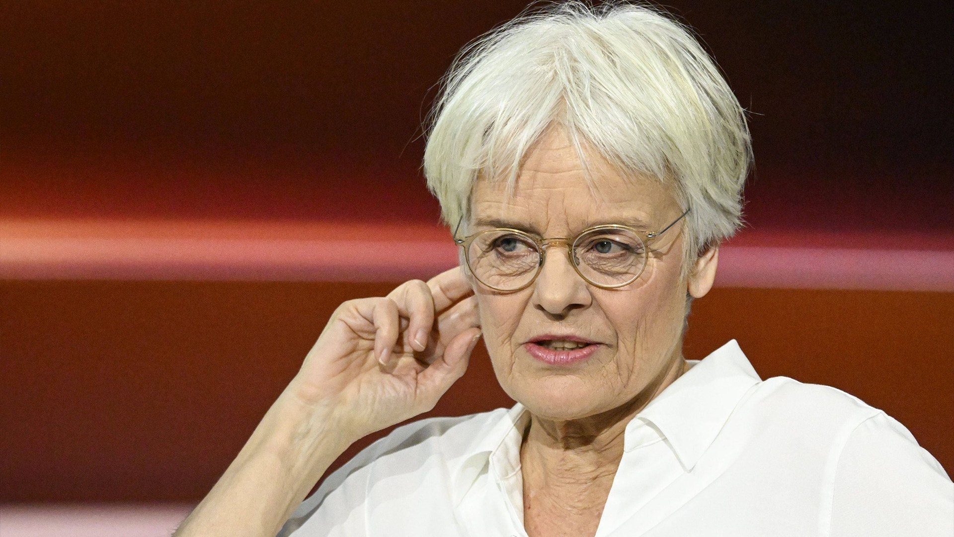 Porträtt av  Ulrike Herrmann med vitt hår och glasögon, klädd i en vit skjorta. Hon lutar sitt huvud mot sin hand i en eftertänksam pose mot en rödbrun bakgrund.