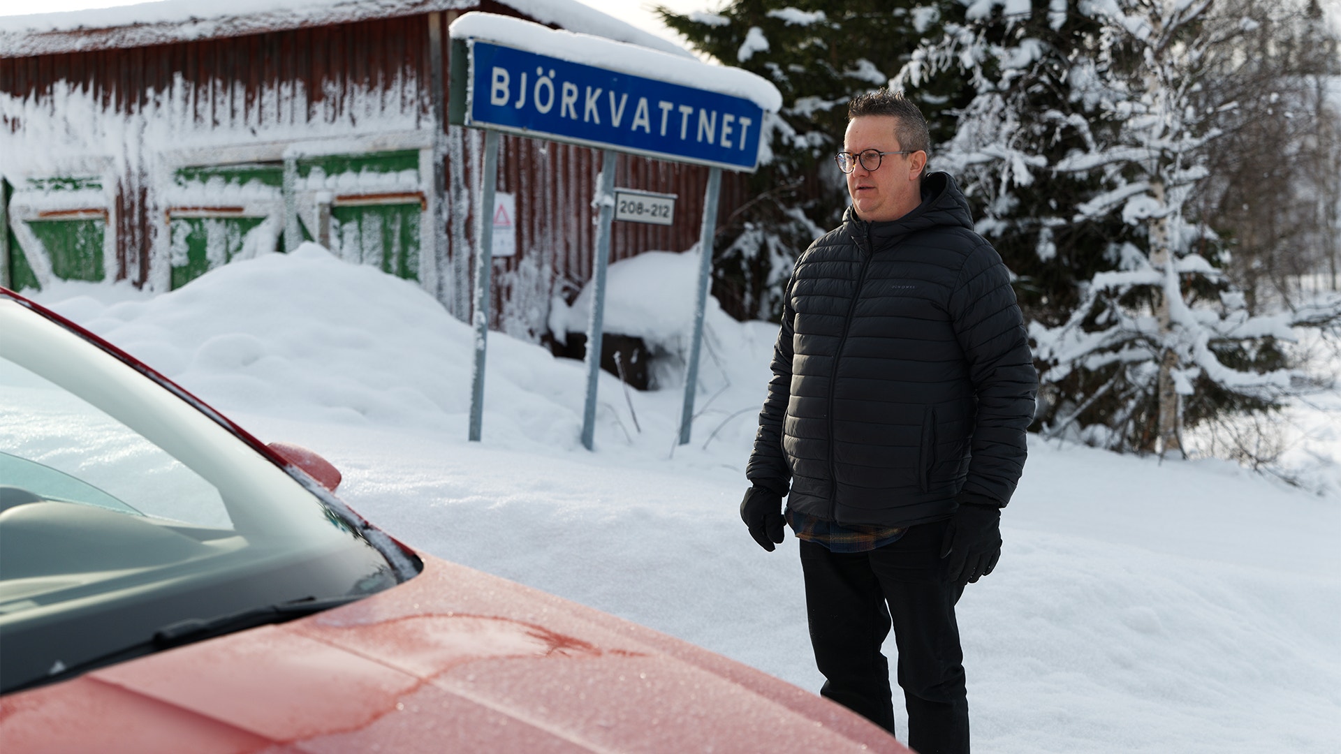 Man i svart jacka står i snölandskap vid vägskylt