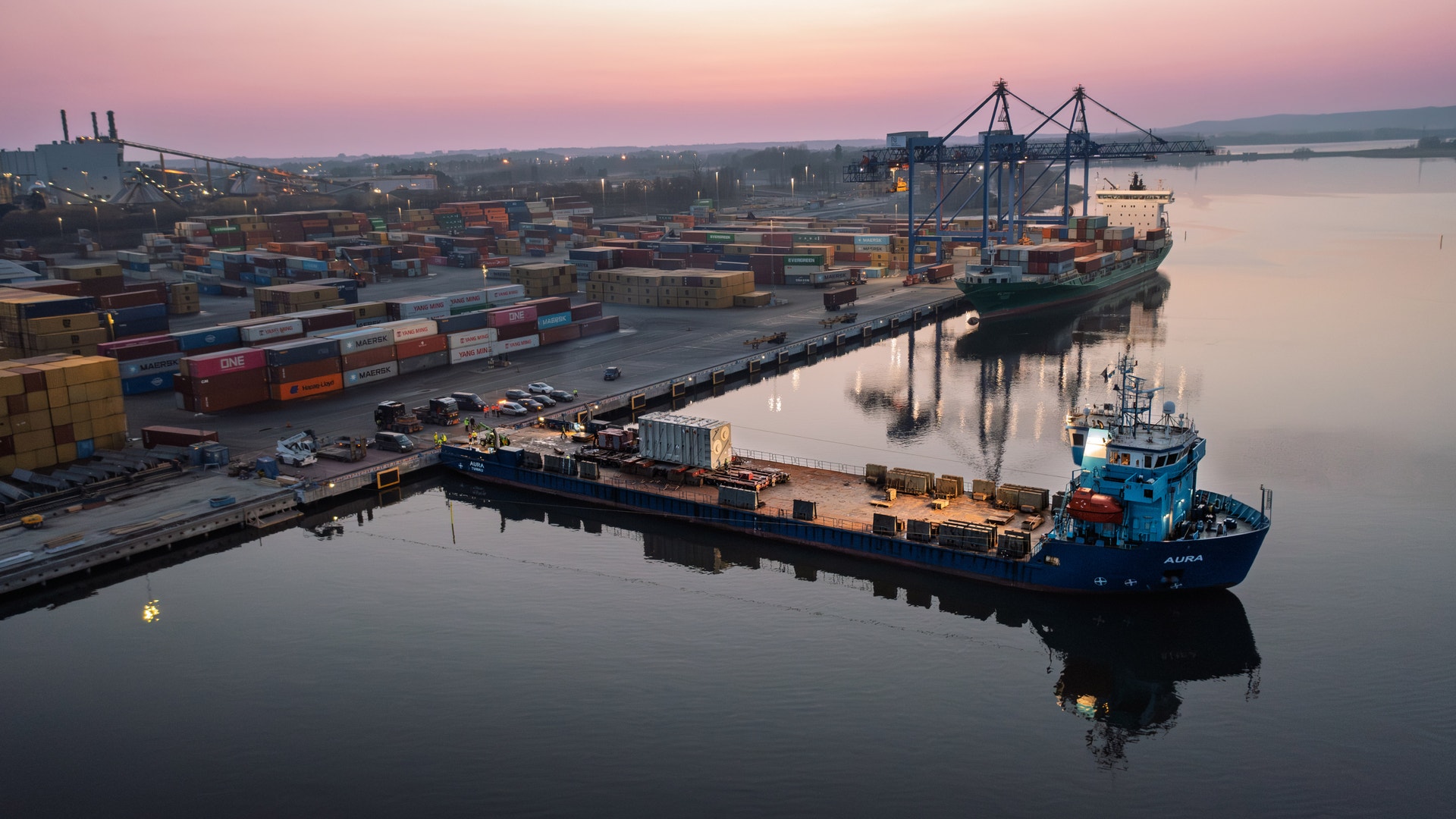 Luftfoto av en containerhamn vid soluppgång. Ett blått fraktfartyg ligger dockat vid kajen omgiven av staplade containrar och en stor containerlyft-kran. Vattnet är lugnt och speglande.