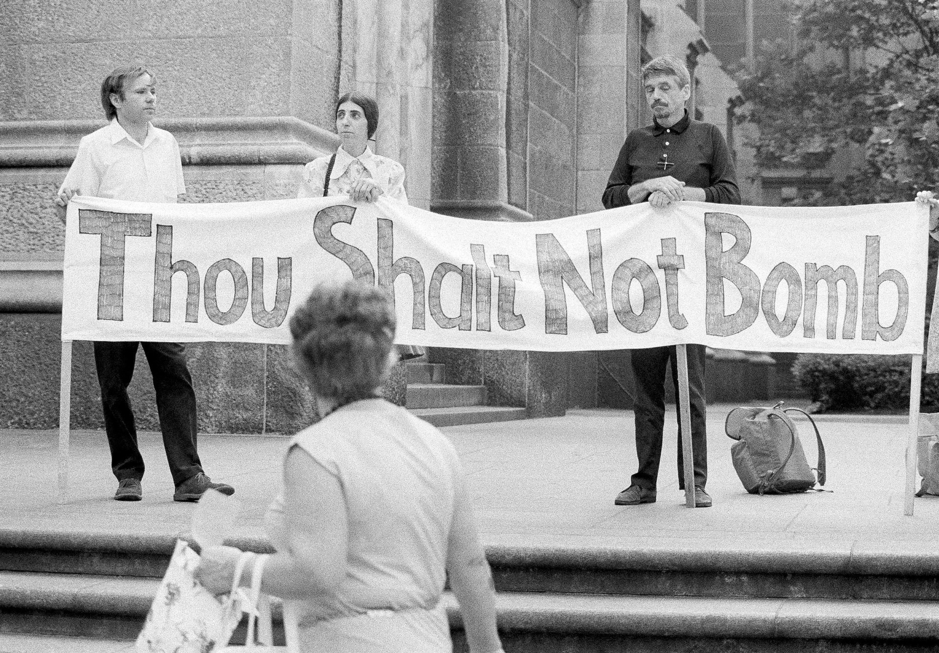 Svartvit fotografi från en fredsdemonstation. Tre personer håller upp en banner med texten "Thou Shall Not Bomb" framför en byggnad. En person i förgrunden tittar på bannern.