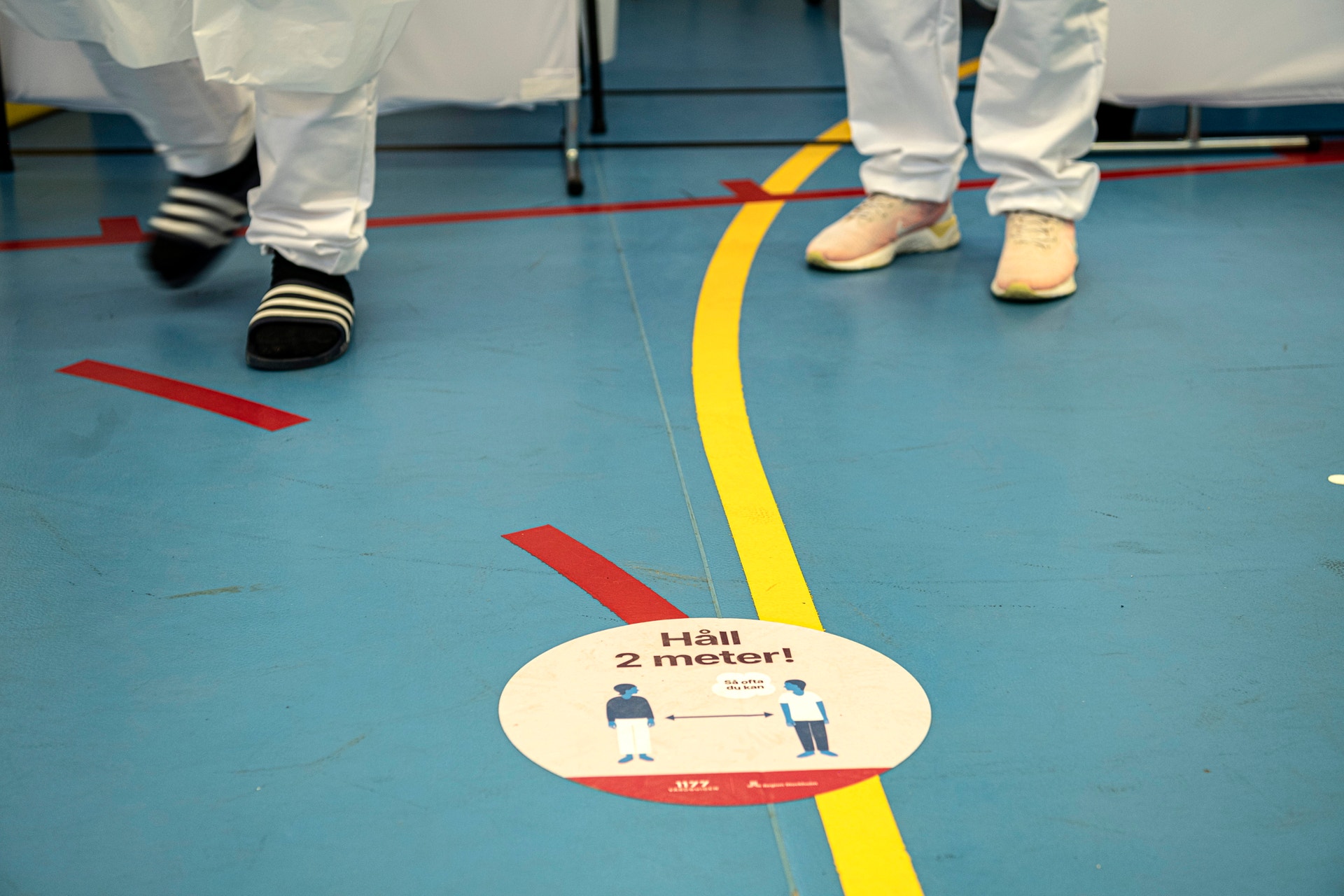Golv markerat med färgade linjer och ett skyltkort som visar instruktionen 'Håll 2 meter' avstånd. Personer i vita kläder står på golvet.