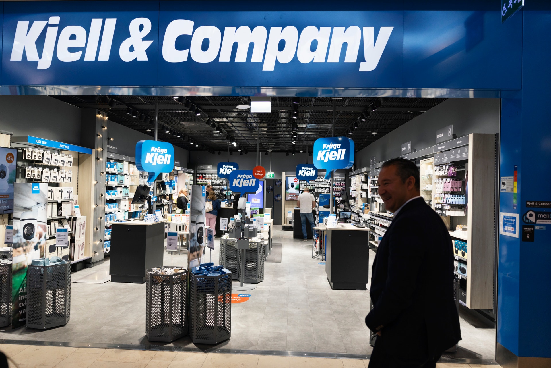 Interiör från en Kjell & Company-butik med blå skyltning, hyllor med elektronikprodukter och en man i mörk kostym som går genom butiken.