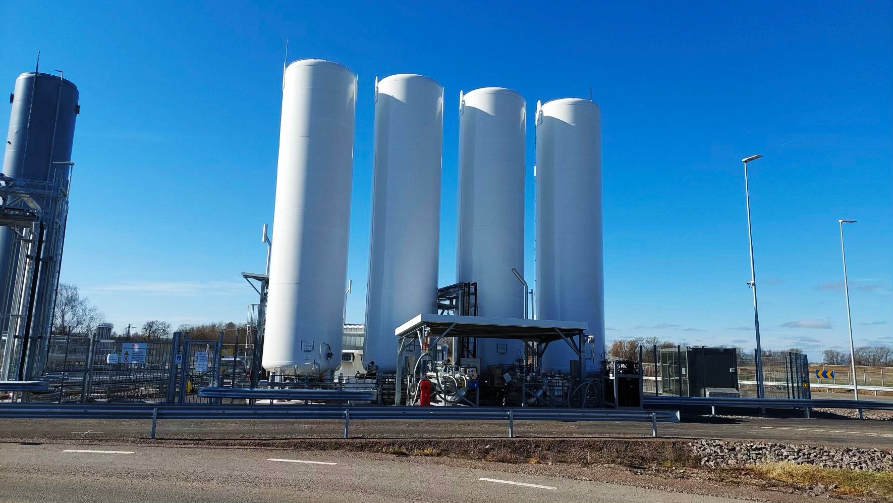 Fyra stora vita cylindriska lagringstankar för biogas vid en anläggning under blå himmel.