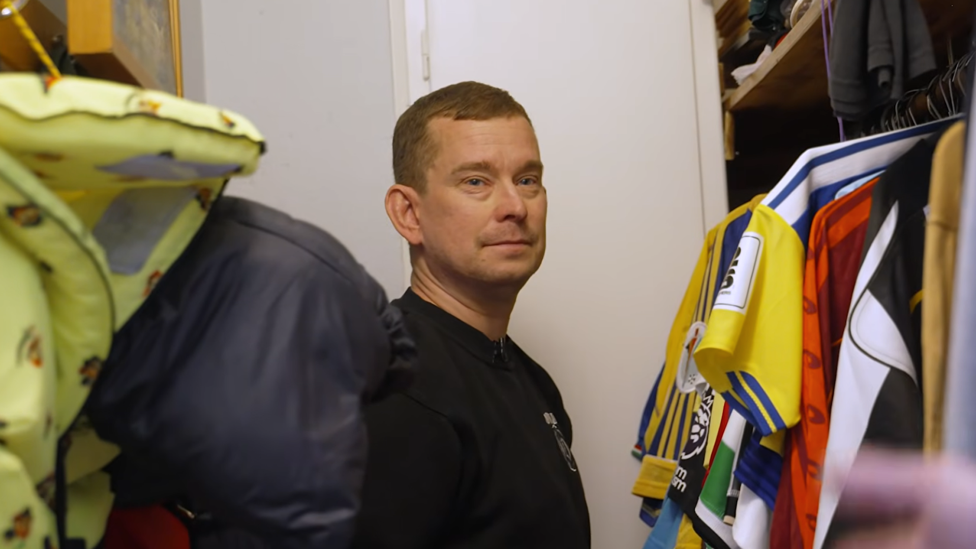 En man i svart tröja står omgiven av färgglada kläder och tröjor som hänger omkring honom i ett klädesrum.