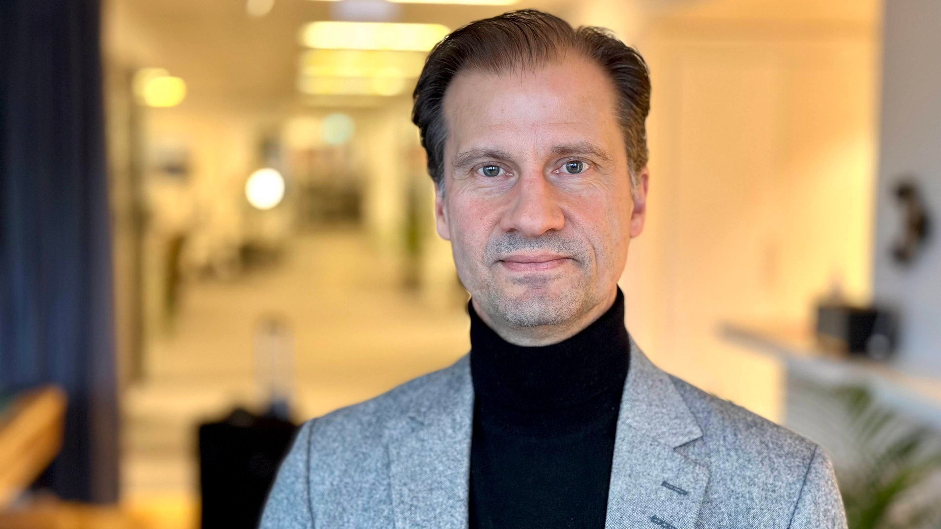 Porträtt av en man i grå kavaj och svart polotröja framför en suddig kontorsinteriör.