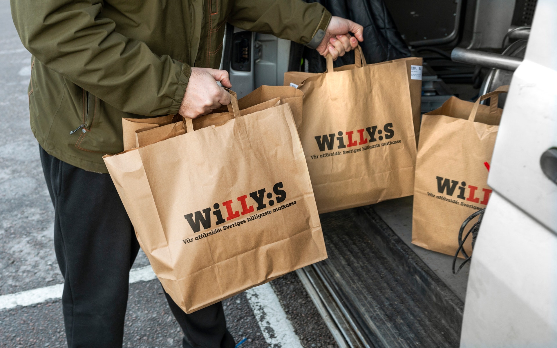En person håller tre papperspåsar märkta WILLY:S framför en öppen bildörr.