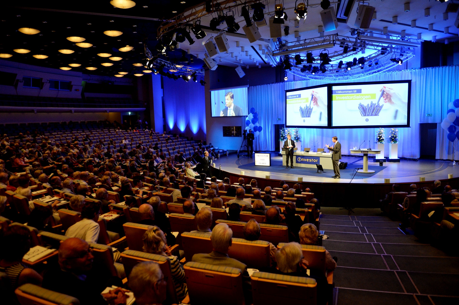 Fullsatt konferenssal där publiken sitter vända mot en scen. På scenen står två personer framför stora skärmar.