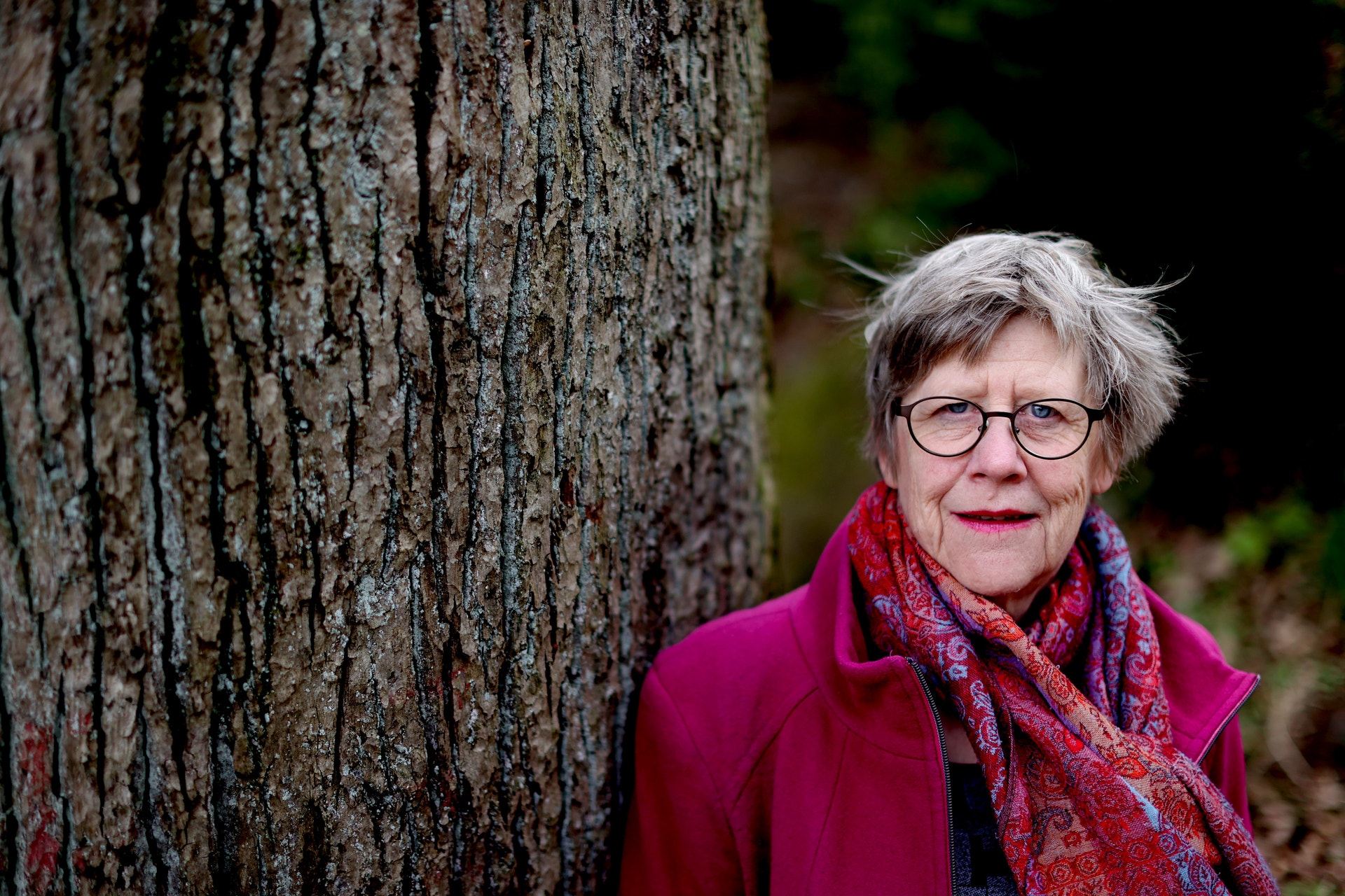 Äldre kvinna med kort grått hår och glasögon, klädd i rosa jacka och röd mönstrad halsduk, står utomhus intill en trädstam.