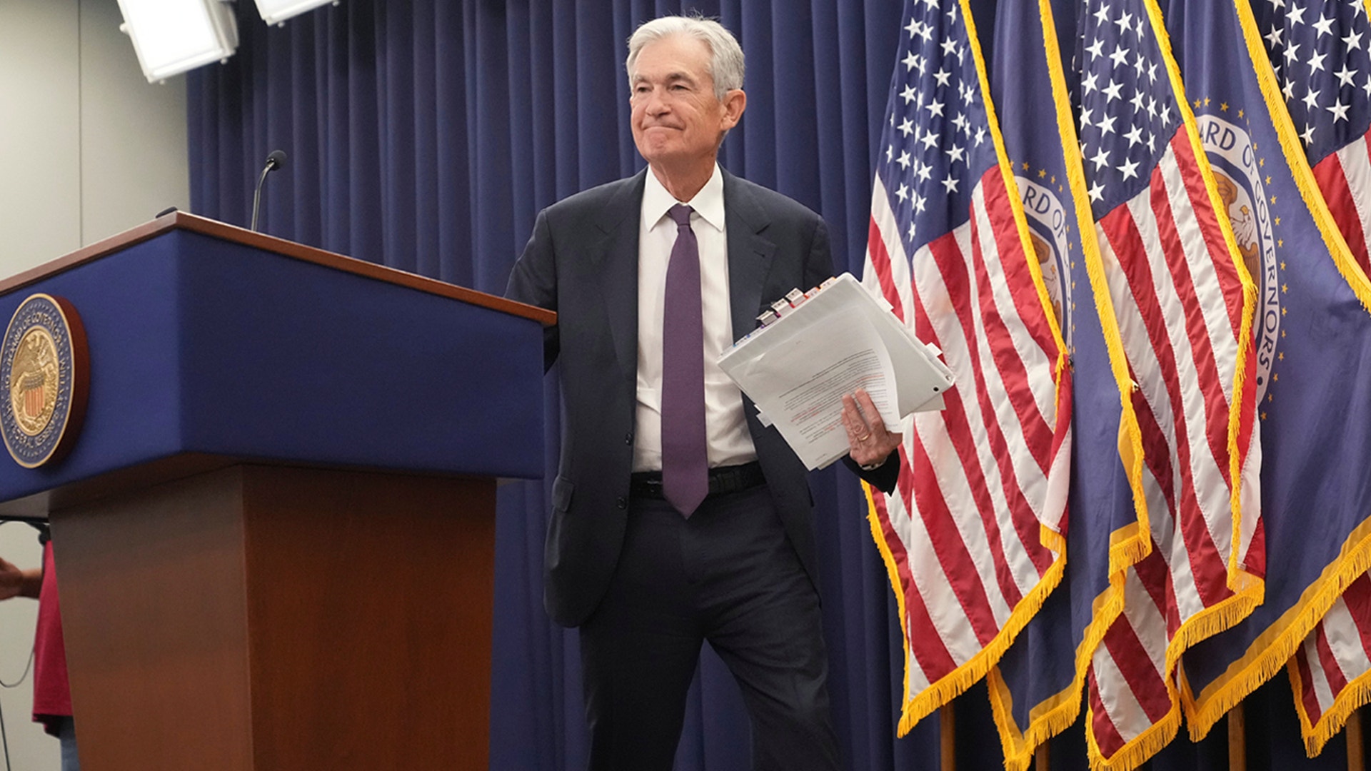 Man i mörk kostym vid podium med amerikanska flaggor i bakgrunden, håller papper i handen.