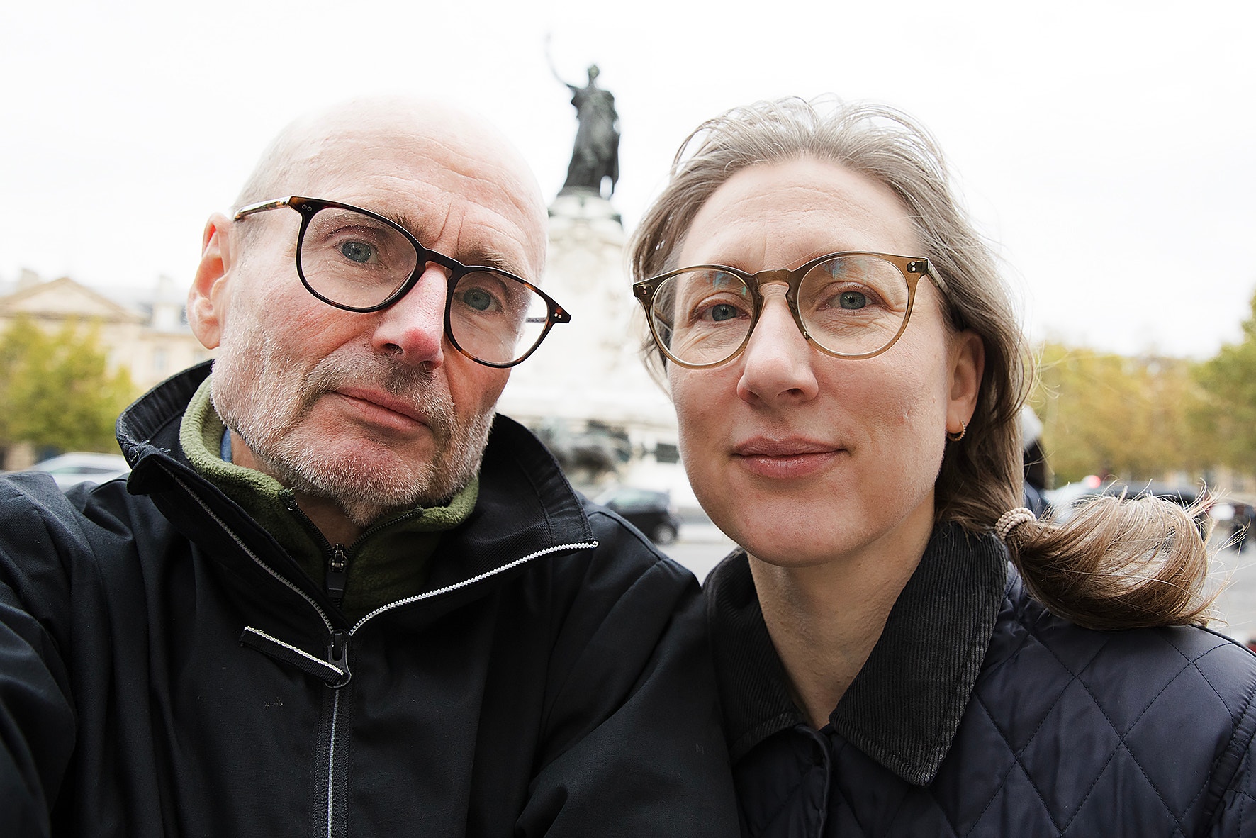 Selfie av en man och en kvinna med glasögon och mörka jackor. Bakom dem syns en staty på en hög sockel utomhus.