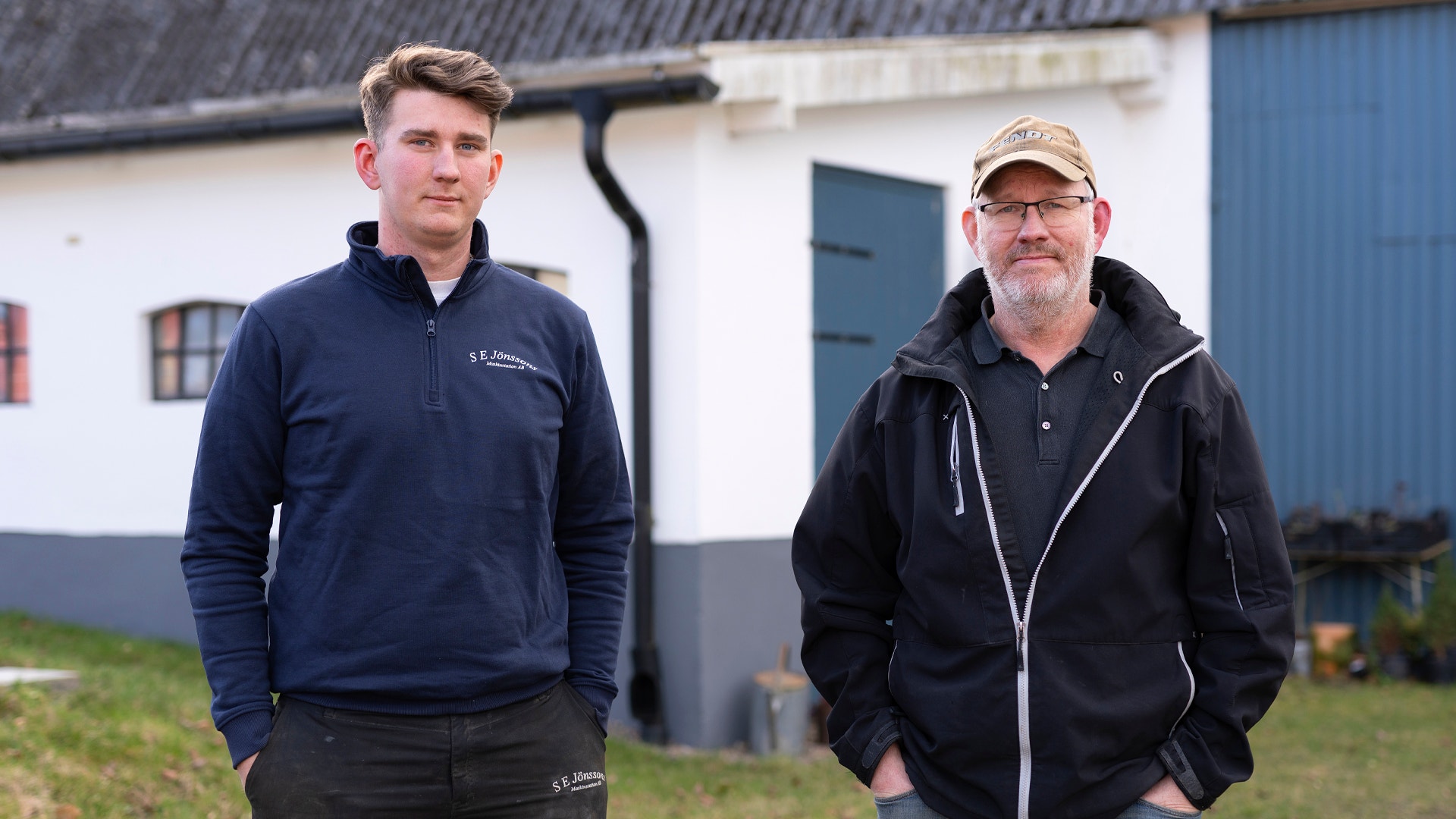 Två män står framför en vit byggnad med blå dörr. Den vänstra mannen bär en marinblå tröja och den högra mannen bär en svart jacka och keps.