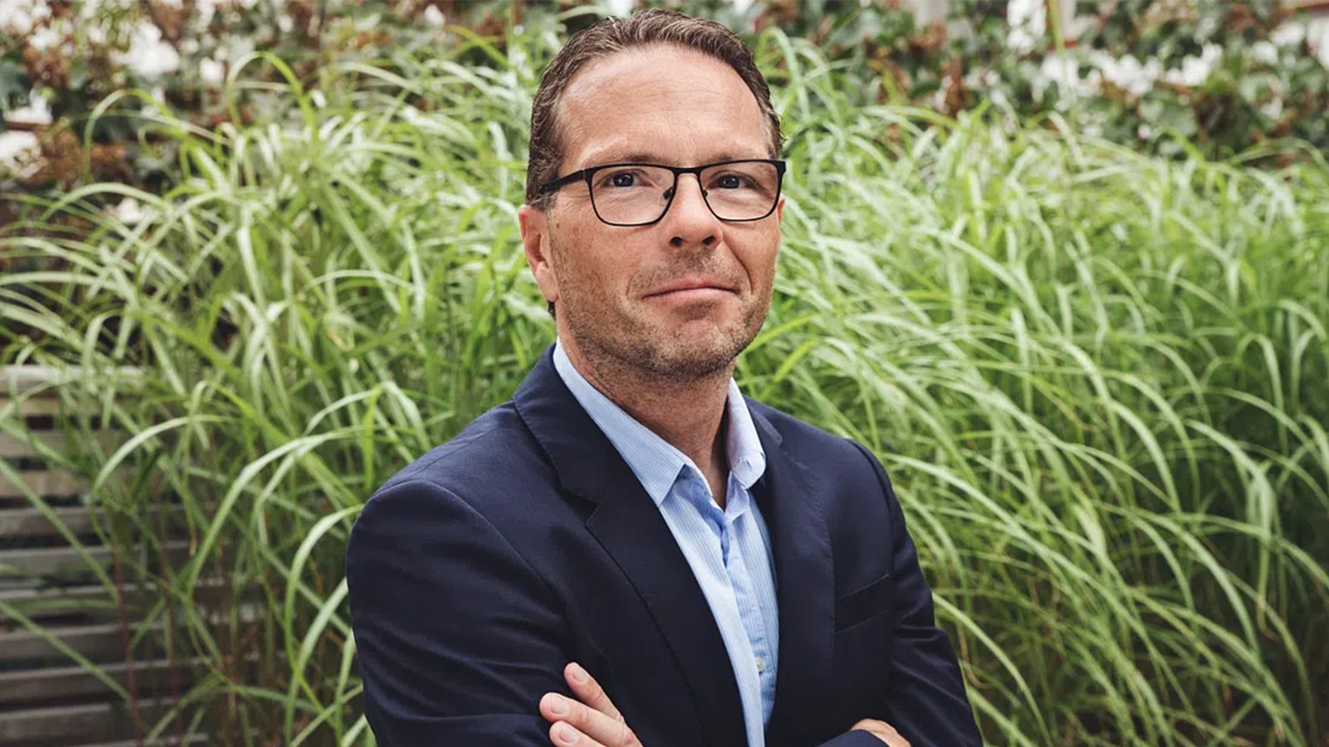 Porträtt av en man med glasögon och mörkblå kavaj framför grön växtlighet.