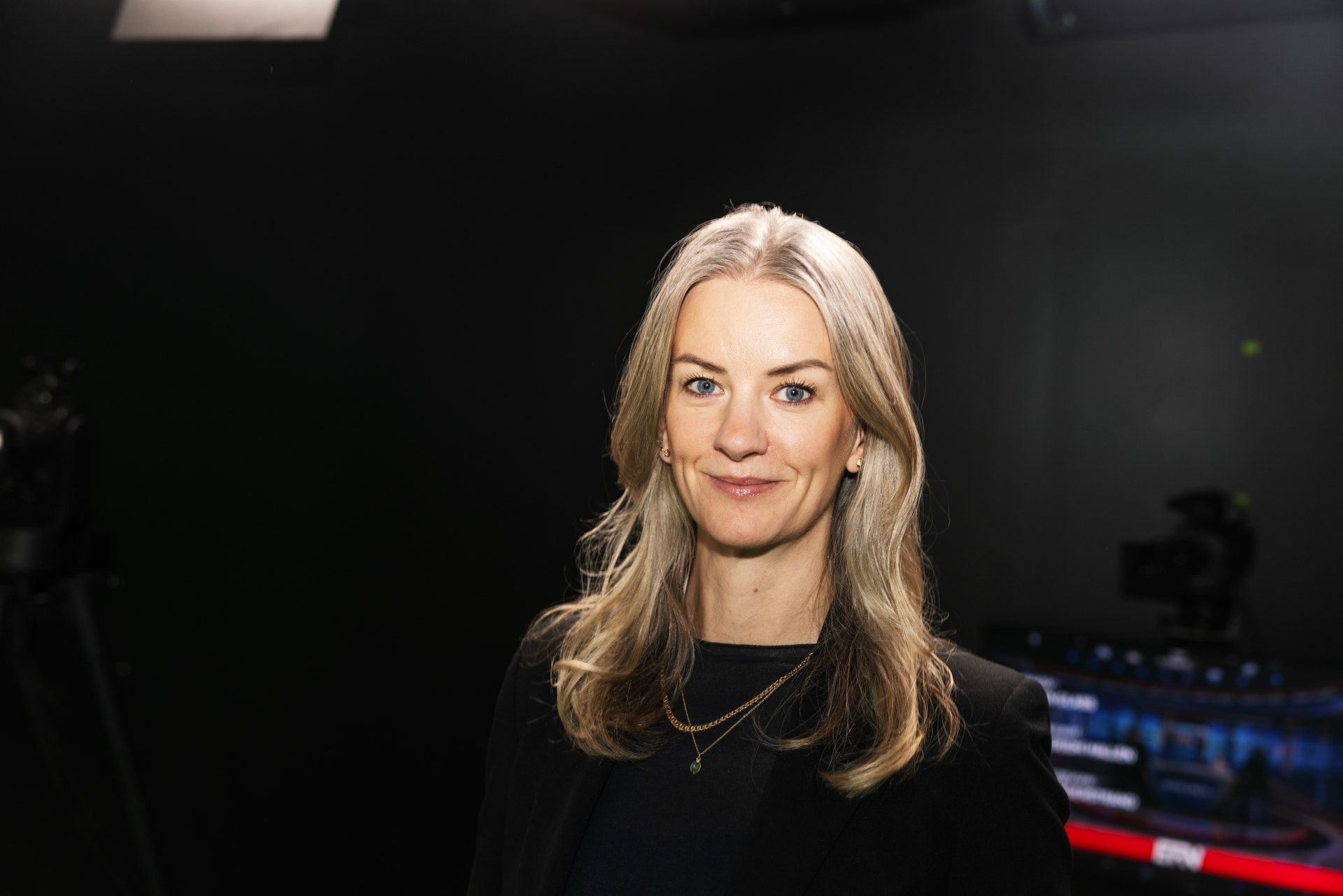 Porträtt av en leende kvinna med långt ljust hår, klädd i svart, i en mörk tv-studio.