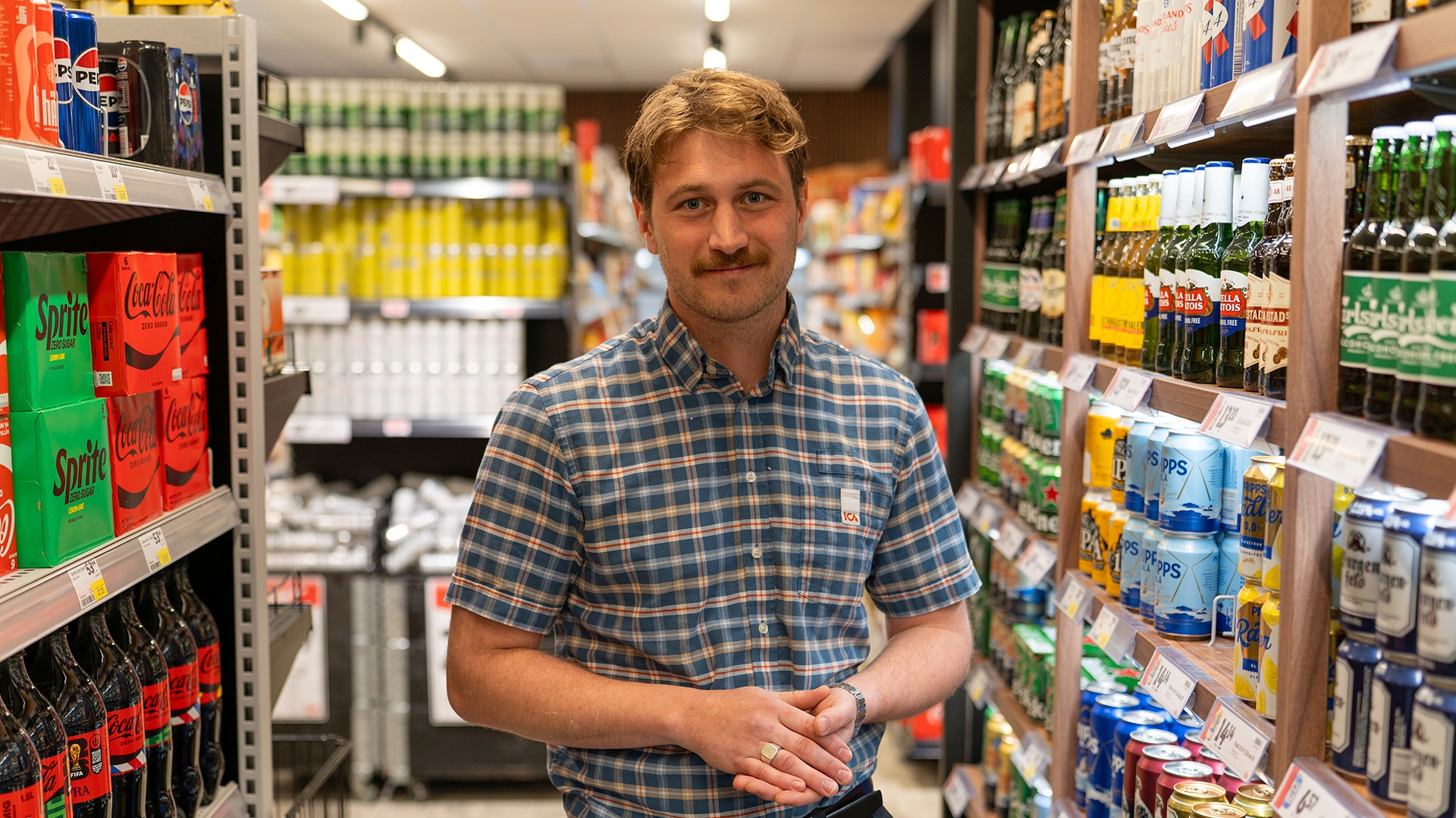 En man i blå rutig skjorta står i en matbutik omgiven av hyllor med drycker och matvaror.