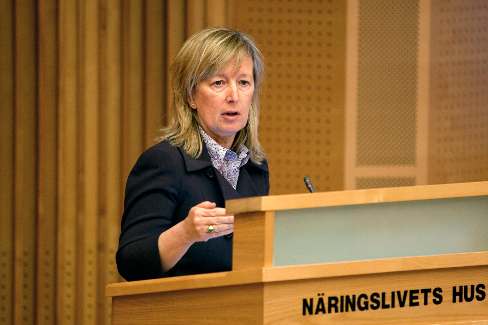 En kvinna talar vid ett podium märkt Näringslivets hus. Hon bär en svart blazer och gestikulerar medan hon presenterar.