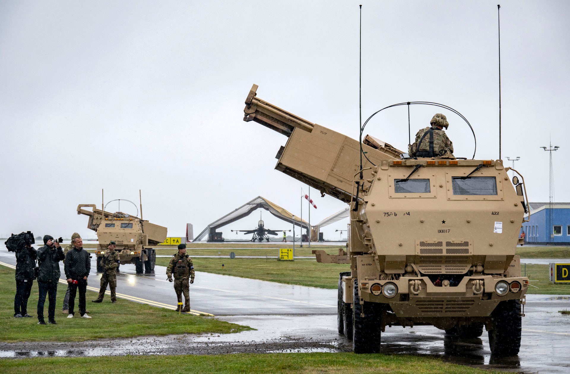 Militärt luftvärnsfordon i sandfärg med vapen monterat på taket, omgivet av soldater på en fuktig militärbas.