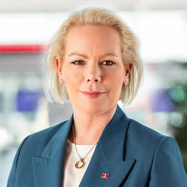 Porträtt av en kvinna med blont hår, iklädd mörkblå kavaj och vit topp, fotograferad mot en suddig bakgrund.