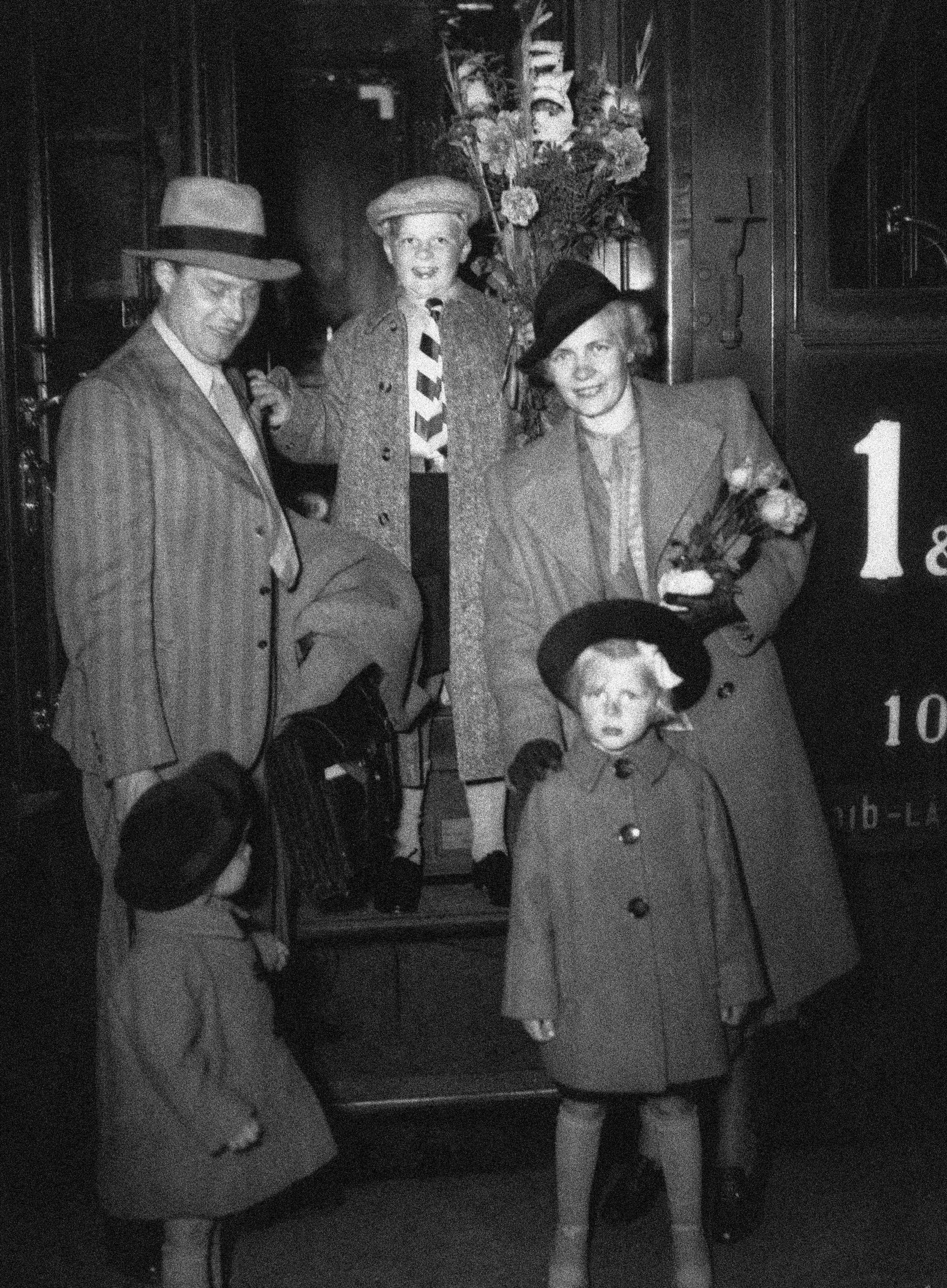 Svartvit fotografi från 1940-talet som visar tre vuxna och ett barn klädda i formella kläder och hattar som står vid en dörr.