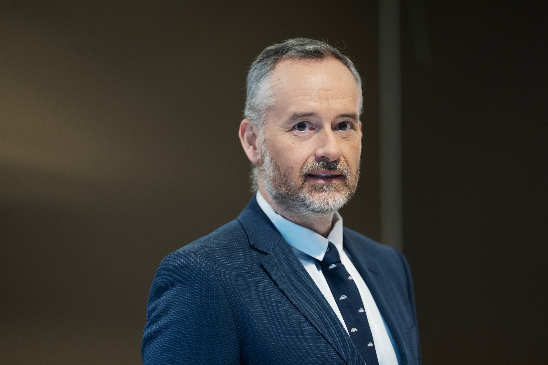 Porträtt av en man i mörkblå kostym och vit skjorta mot en neutral bakgrund.