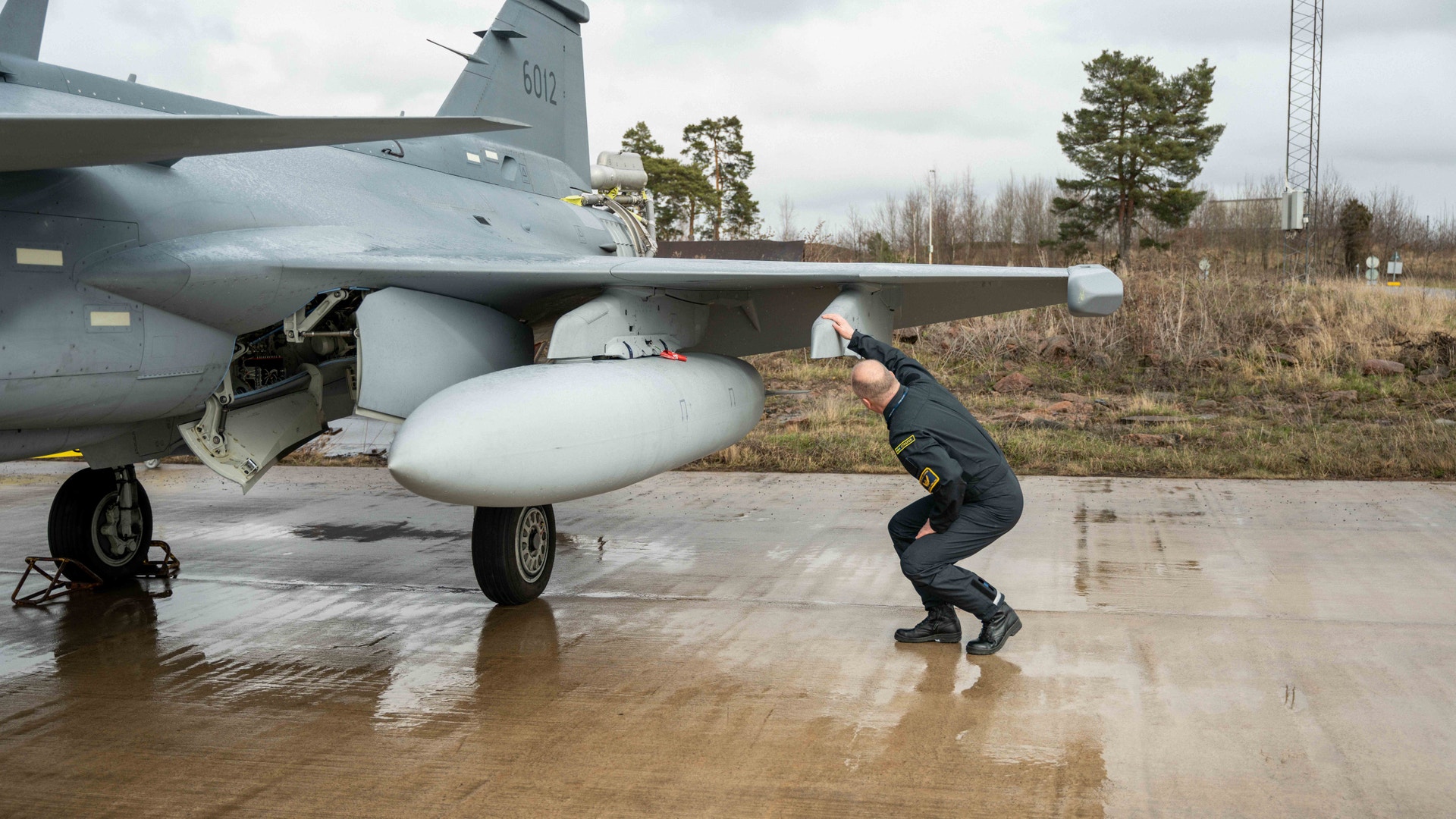 En person inspekterar underredet på ett militärt stridsflygplan som står parkerat på en våt betongplatta.