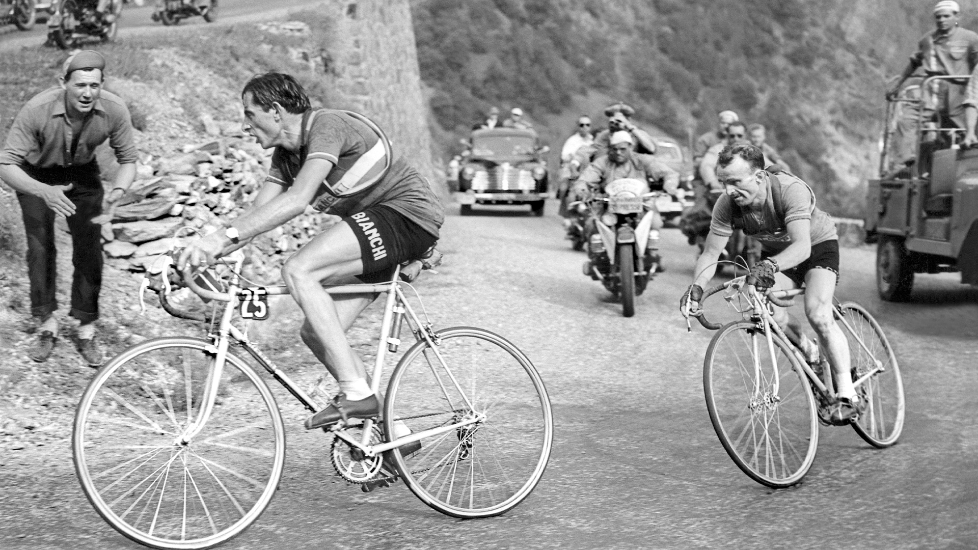 Svartvit fotografi från en cykelkappning på en bergväg med cyklister och åskådare längs vägen.