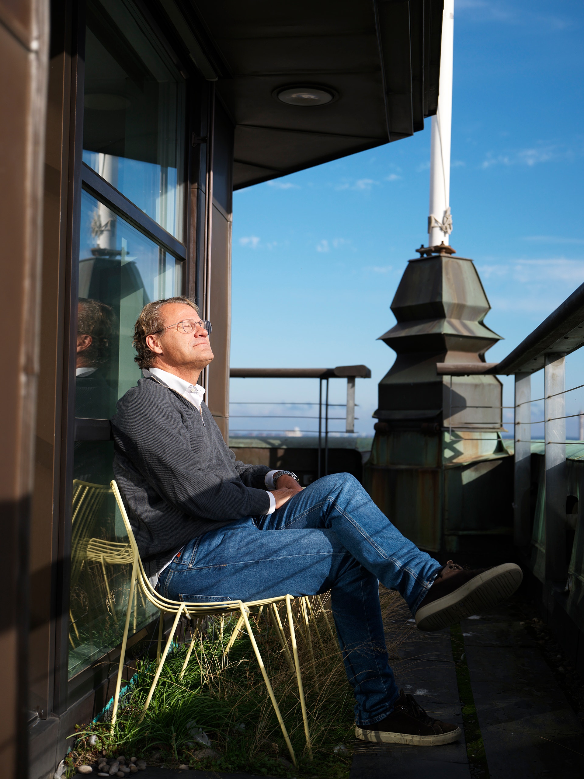 En man sitter på en stol på en balkong och tittar upp mot himlen i solsken.